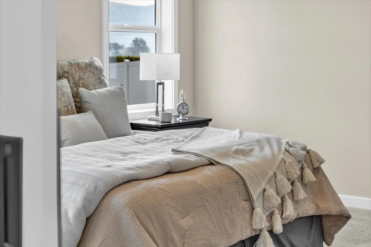 A neatly made bed with cream, white, and beige bedding, pillows, and a blanket, next to a window with a black nightstand holding a lamp, clock, and decorative item, in a bright bedroom.