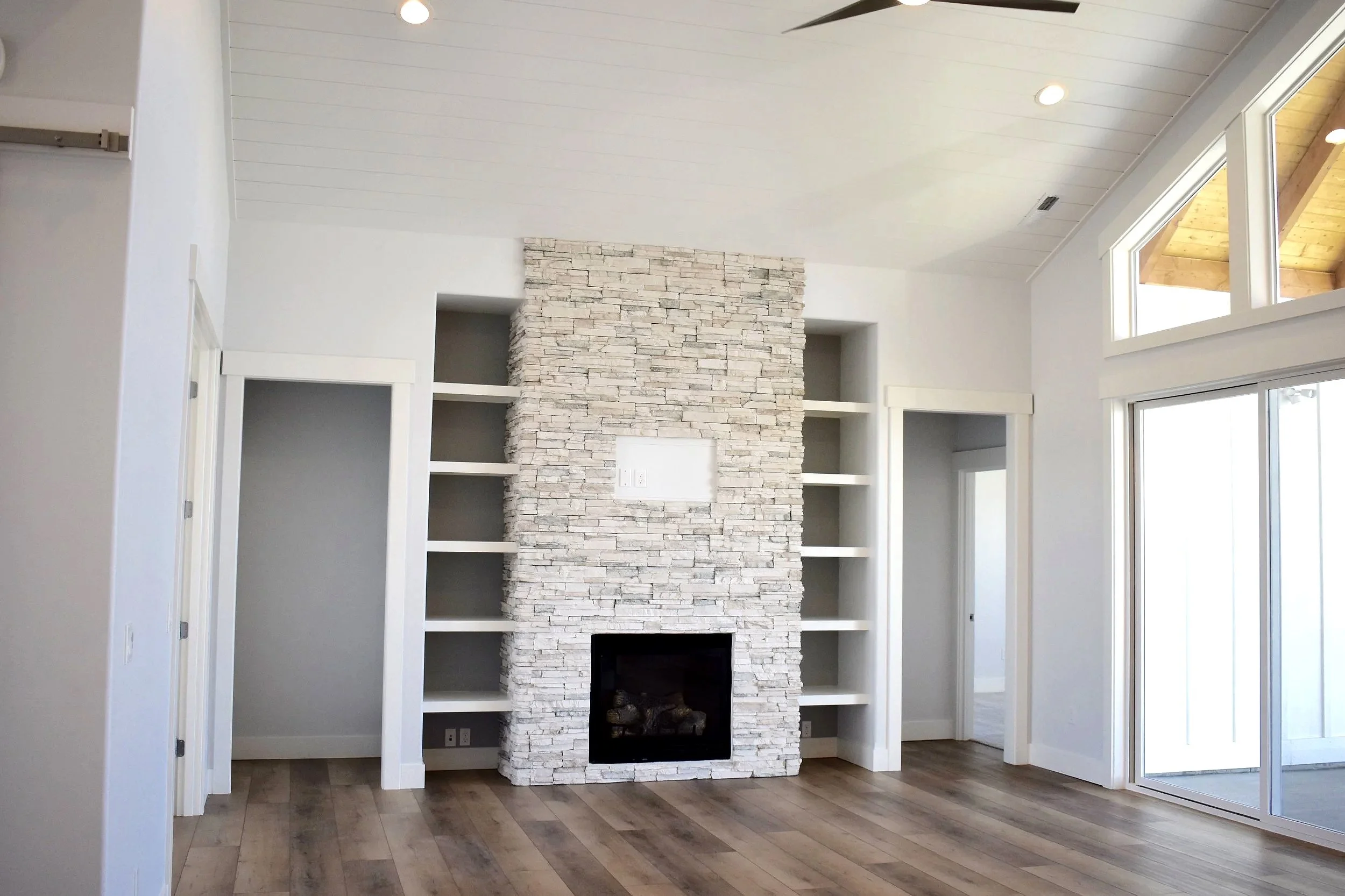 Living room with a white stone fireplace, built-in shelves on either side, large sliding glass door, and vaulted ceiling with recessed lighting.