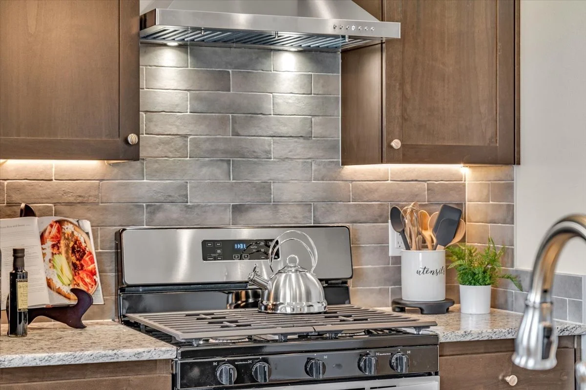 Kitchen stove with stainless steel kettle, gray tiled backsplash, wooden cabinets, and various cooking utensils in a white container and a small potted plant on the counter.