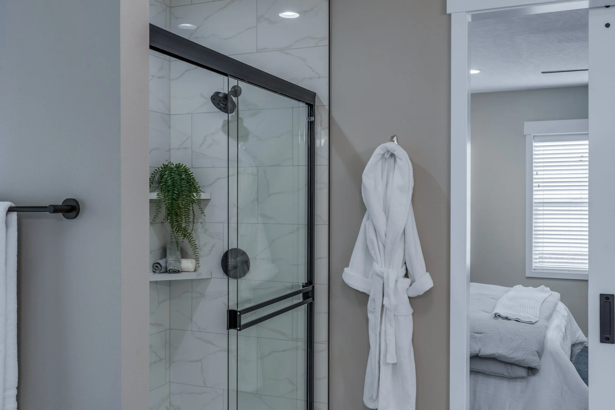 View of a modern bathroom with a glass shower enclosure, white marble-like tile inside, a white bathrobe hanging on a hook outside the shower, and a bedroom visible through an open doorway with a bed and window.