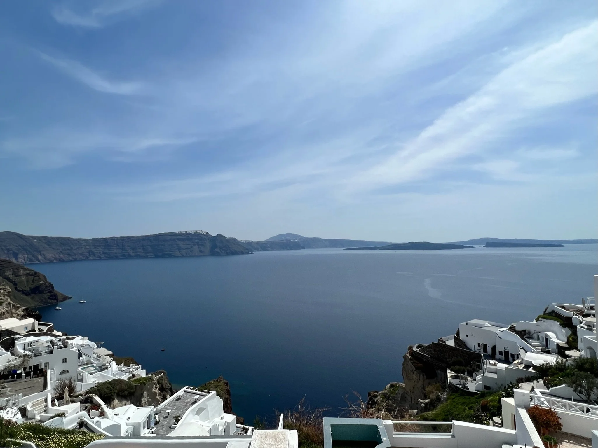A panoramic view of clear blue ocean water with white buildings on cliffs in the foreground and distant islands on the horizon under a partly cloudy sky.