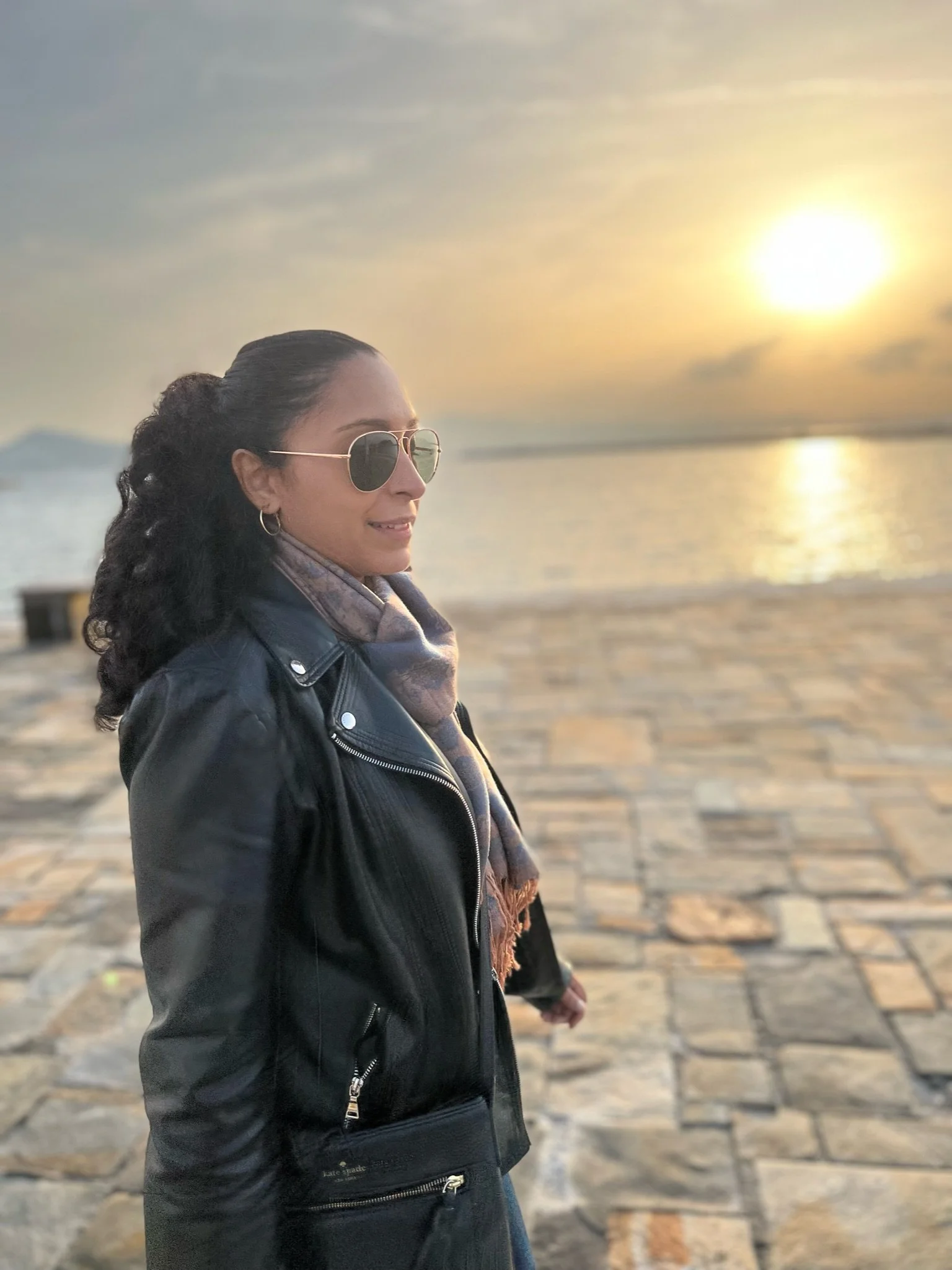 Woman with long curly hair wearing sunglasses and a black leather jacket standing on a stone-paved waterfront at sunset.