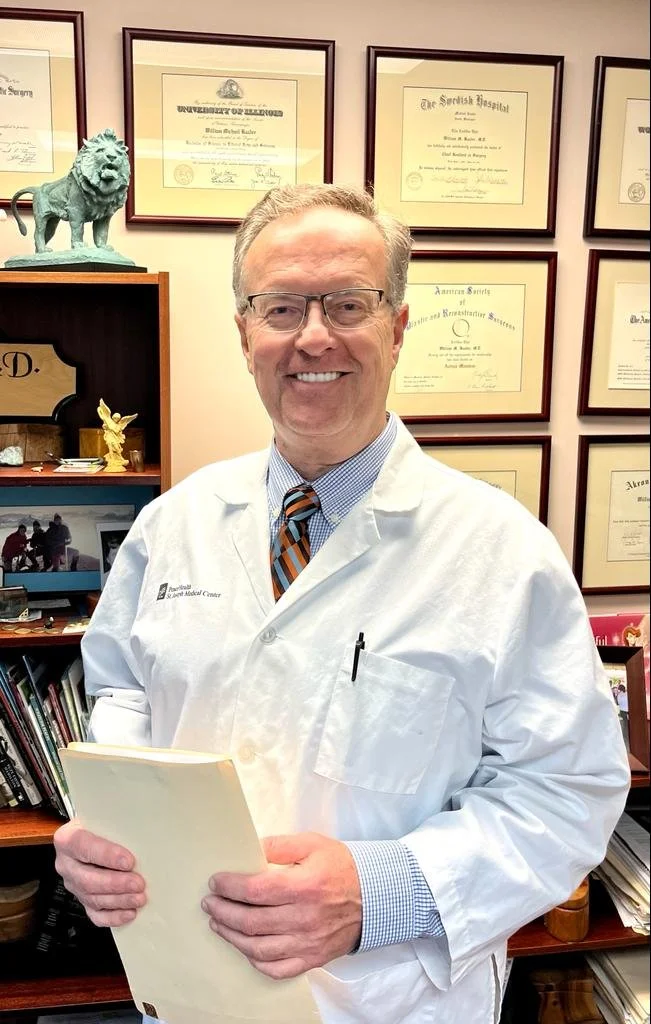 A man in a white medical coat with a blue shirt and striped tie, smiling while holding a folder, standing in an office with framed diplomas and awards on the wall behind him.