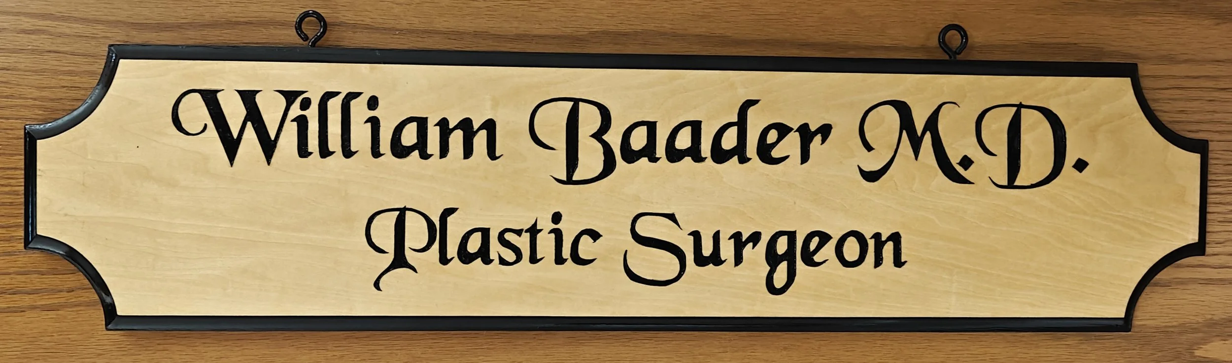 Wooden nameplate with black border reading 'William Baader M.D. Plastic Surgeon' in black ornate letters.
