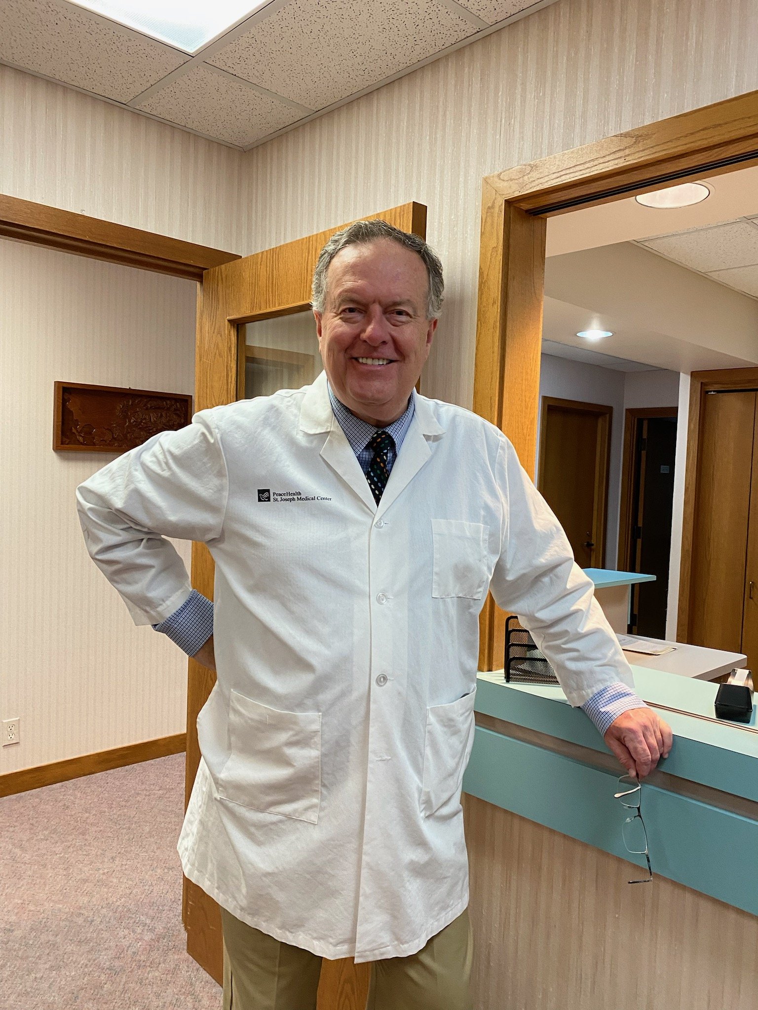 A smiling man in a white lab coat standing at a reception desk in a medical office.