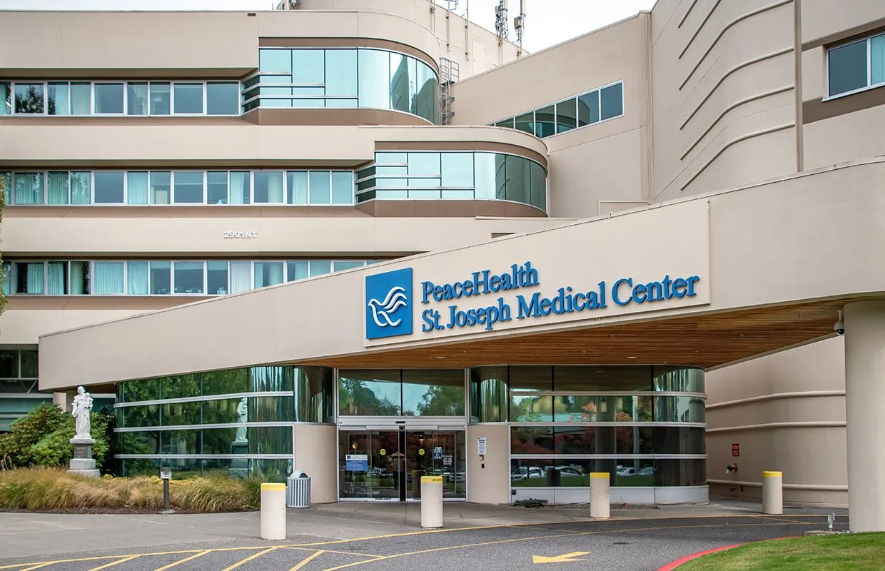 The entrance of PeaceHealth St. Joseph Medical Center, a modern hospital building with large glass windows and a sign displaying its name. The exterior is light-colored with rounded architectural features and some greenery nearby.
