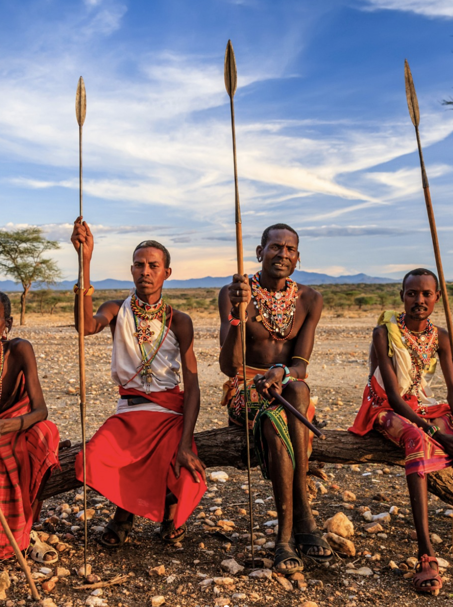 Three Maasai men sitting on a log holding spears, wearing traditional bead necklaces and red shuka skirts, in a savannah landscape with a sunset sky.