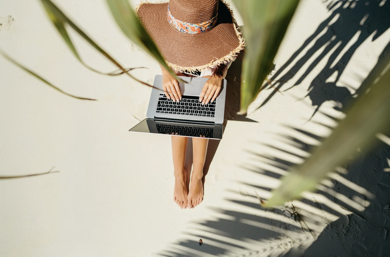 Person barefoot on sandy beach, wearing a large straw hat with a colored band, working on a laptop, with large green palm leaves casting shadows.