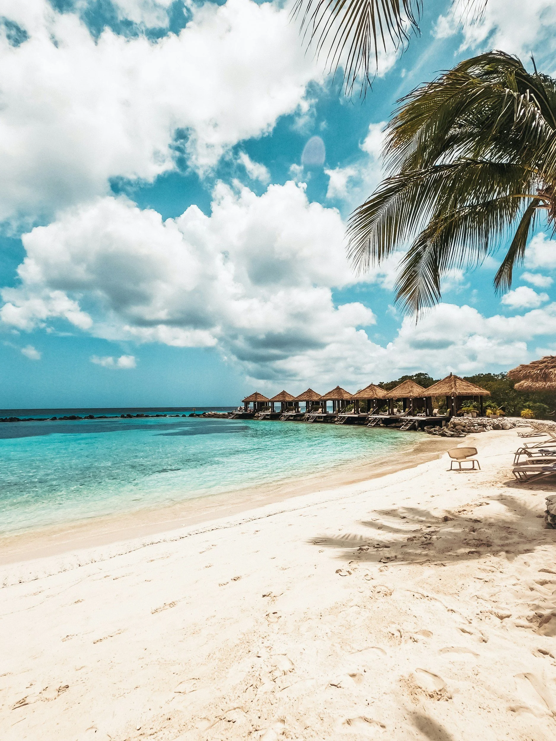 Tropical beach with white sand, turquoise water, overwater bungalows, palm trees, and partly cloudy sky.