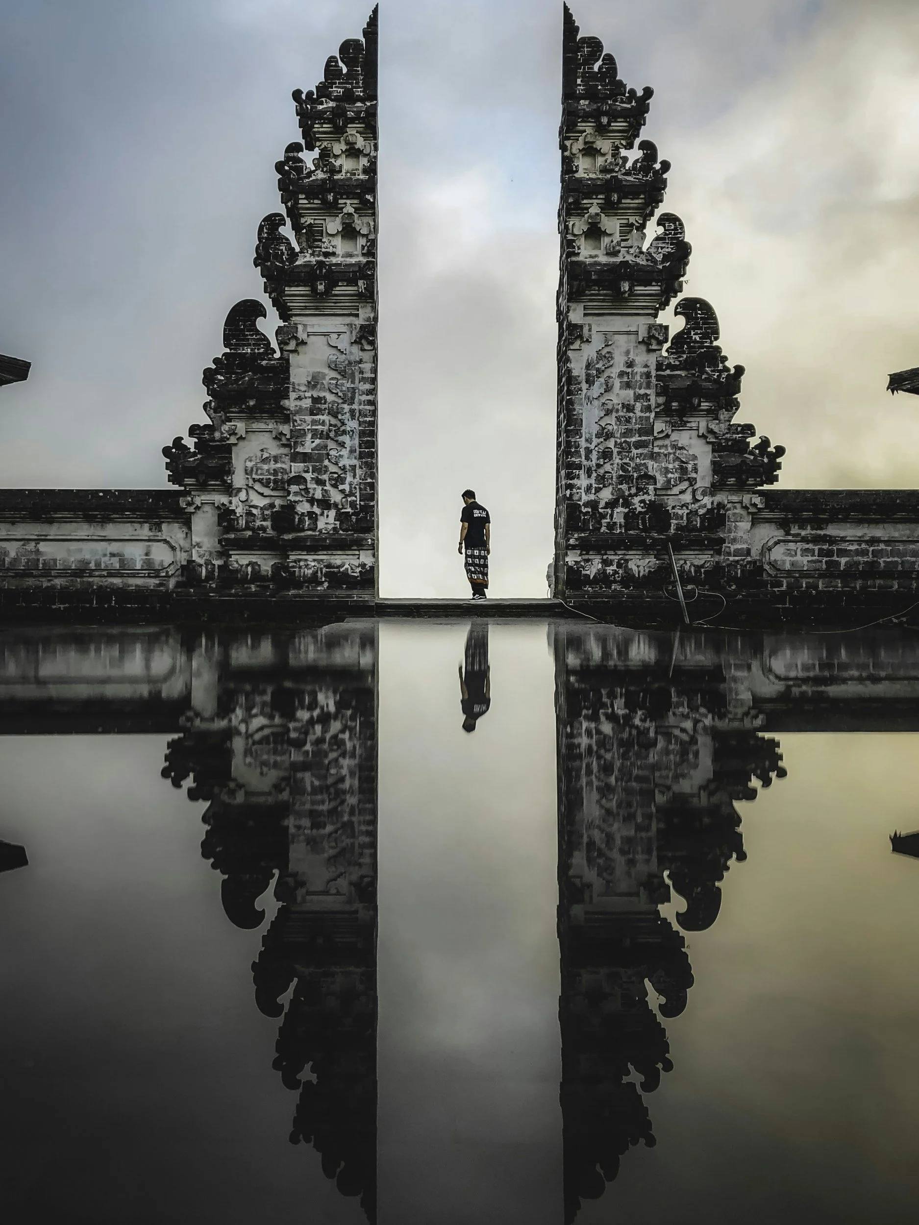 A person standing between two ornate stone gate towers with their reflection in a body of water below. The sky is cloudy and the setting appears to be a traditional Balinese temple entrance.