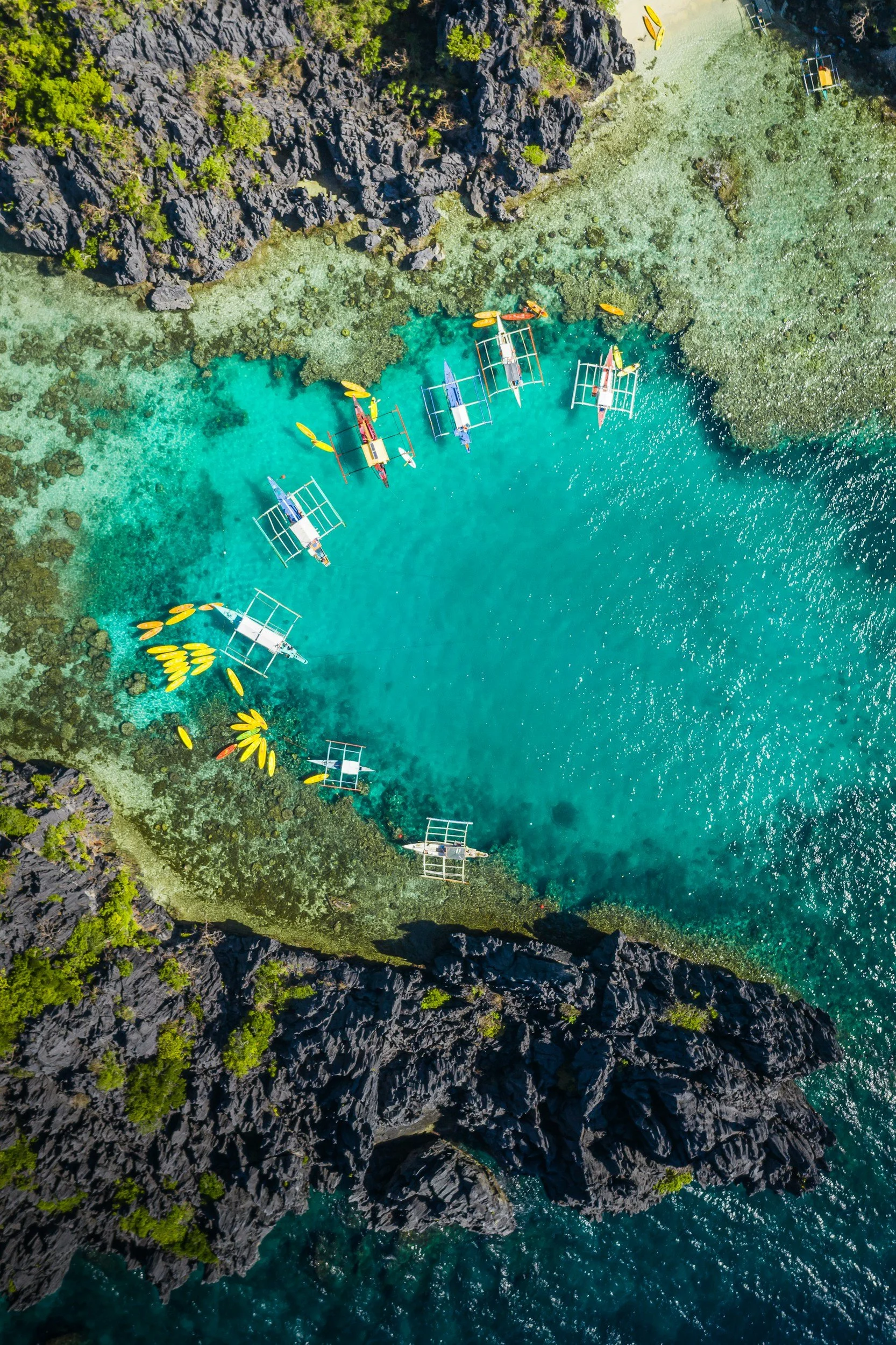 An aerial view of a turquoise coastal area with black rocky cliffs on either side. Several traditional boats with outriggers and kayaks are anchored near the shore. The water is clear, showing coral reefs and seabed underneath.