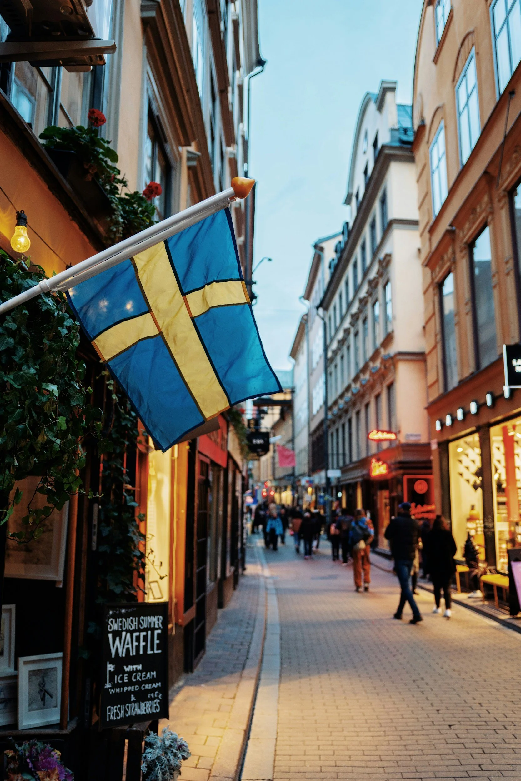 Swedish flag hanging outside a building on a busy city street during evening with shops and pedestrians.