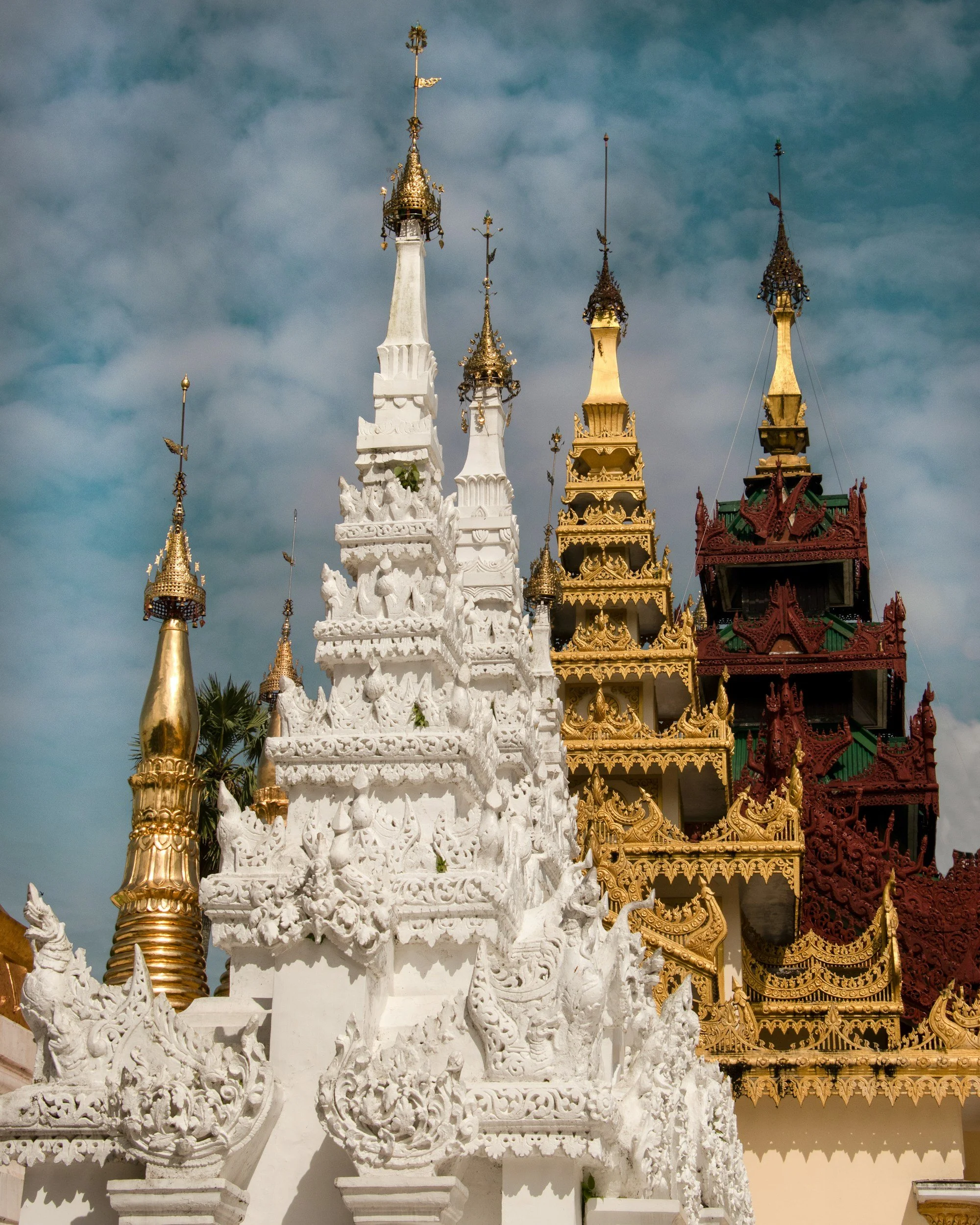 Traditional Thai temple with ornate, tiered roofs and spires, painted white and gold, set against a partly cloudy sky.