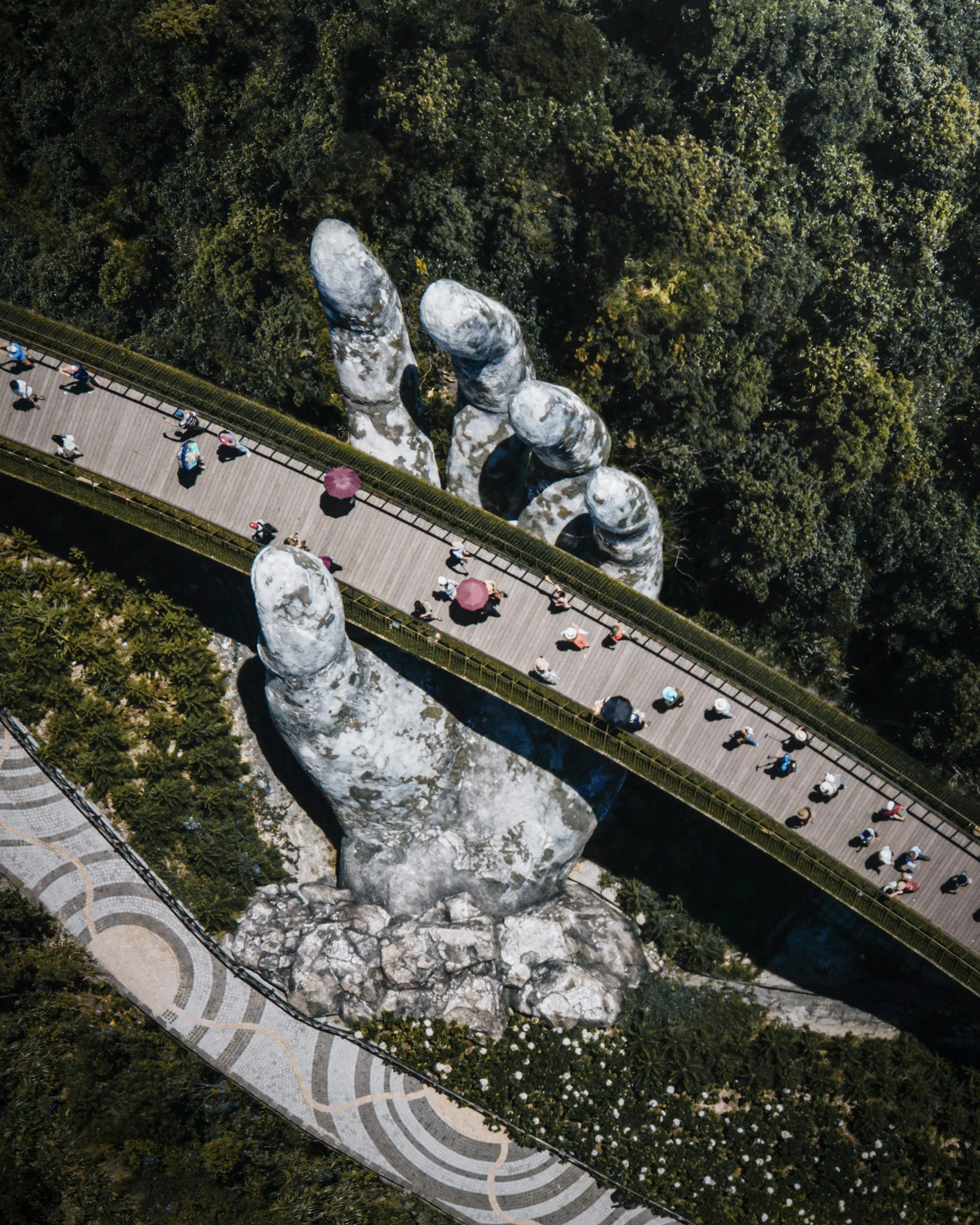 An aerial view of a large hand sculpture emerging from the ground, with a pathway running through the palm and fingers. People are walking along the pathway, some holding umbrellas, surrounded by lush green trees and a patterned walkway.