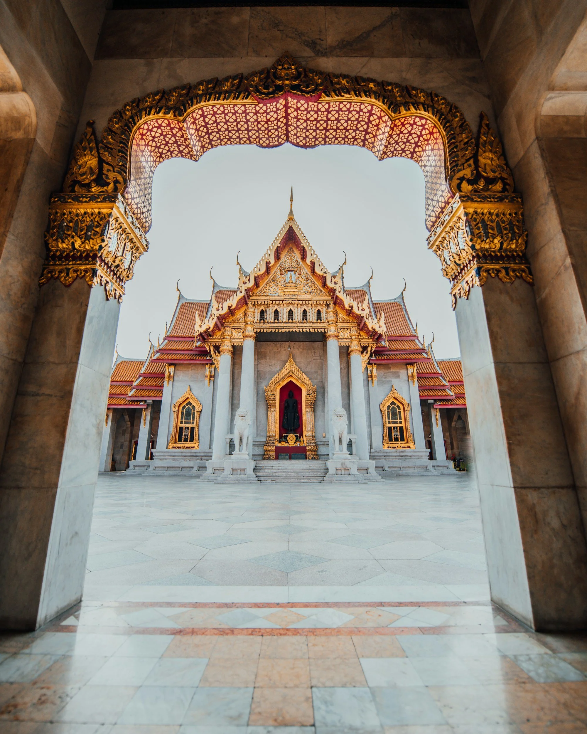 View of a traditional Thai temple with ornate gold detailing and a red-tiled roof, seen through an intricately decorated archway.