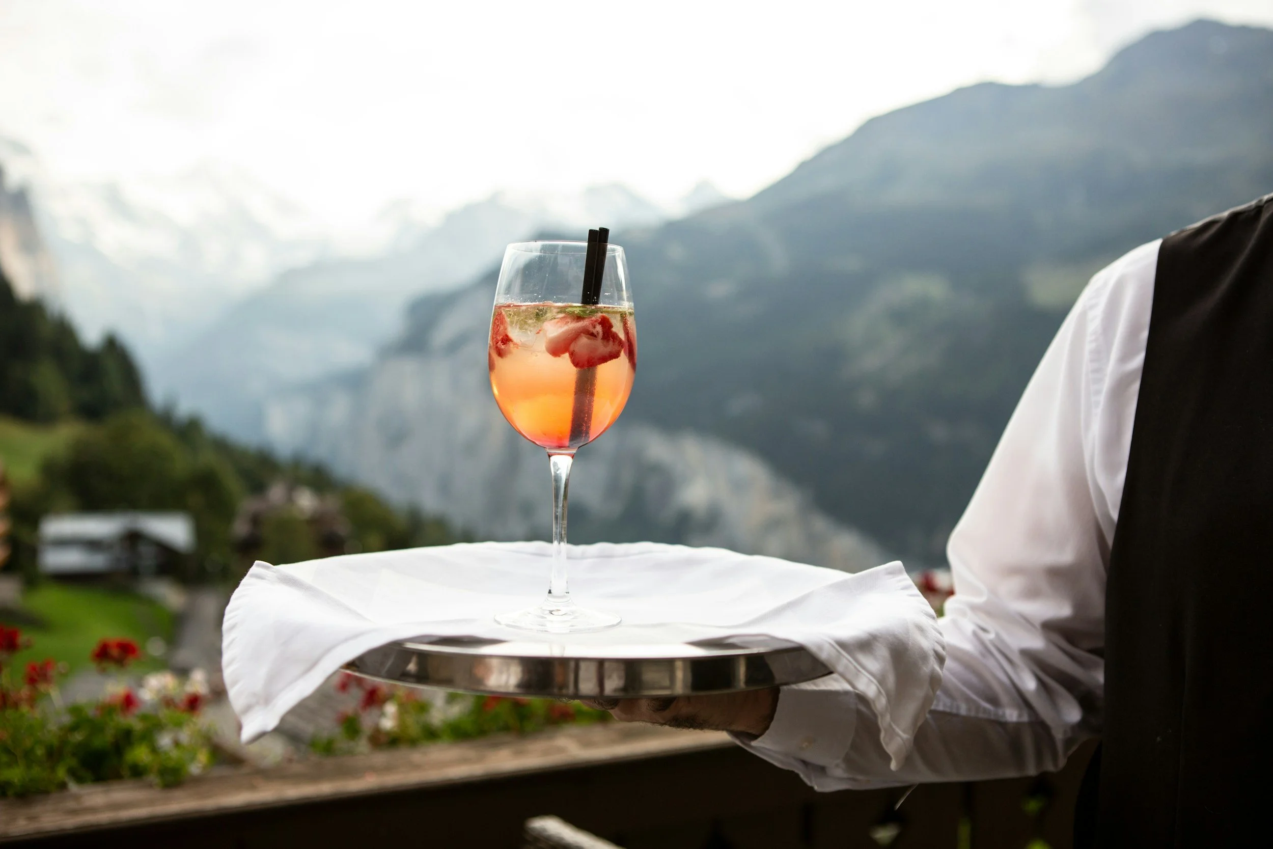 A waiter holds a silver tray with a white cloth, on which sits a cocktail glass filled with a pinkish drink containing berries and a black straw, with a scenic mountain backdrop.