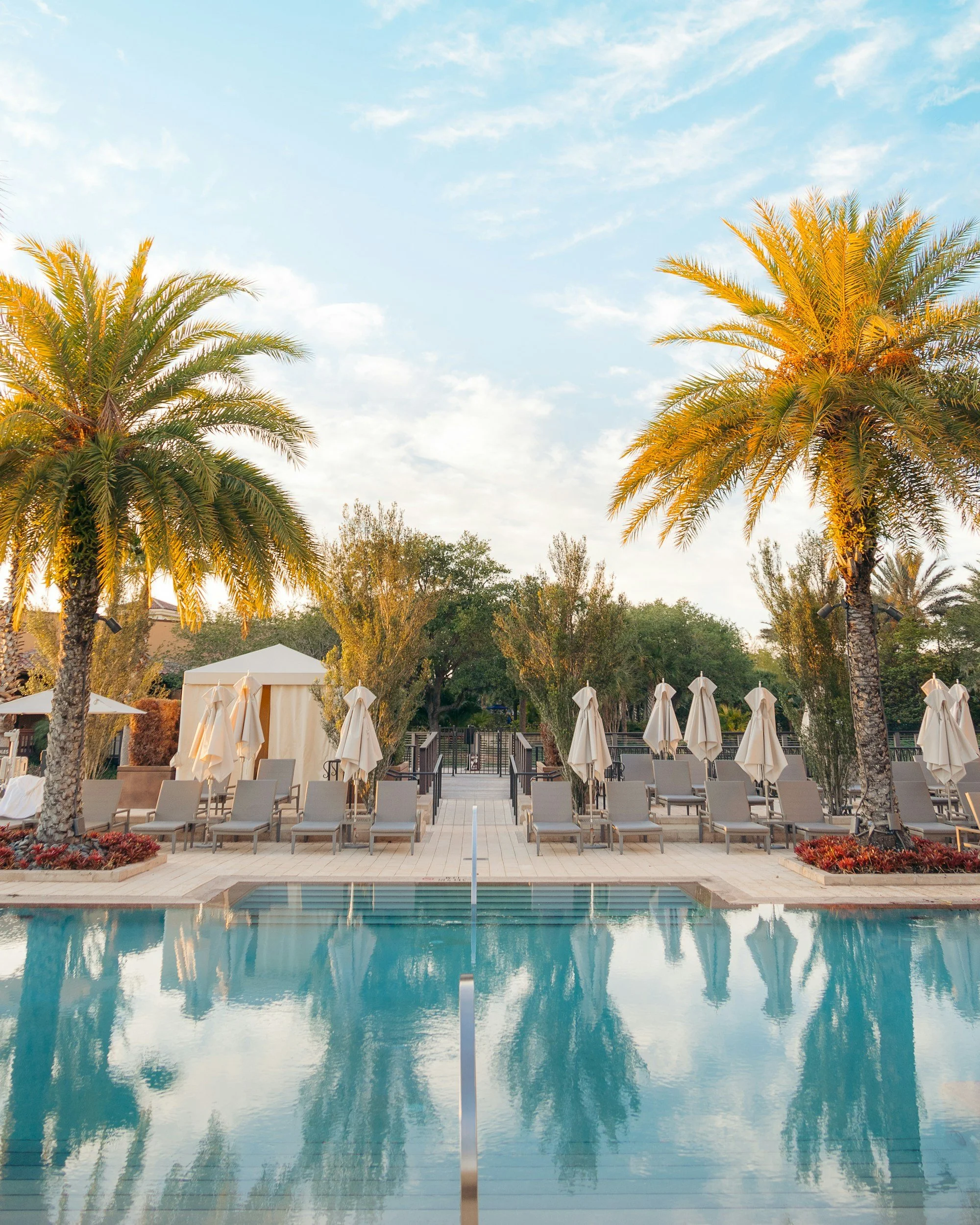 Poolside area with lounge chairs, umbrellas, palm trees, and a cloudy sky.