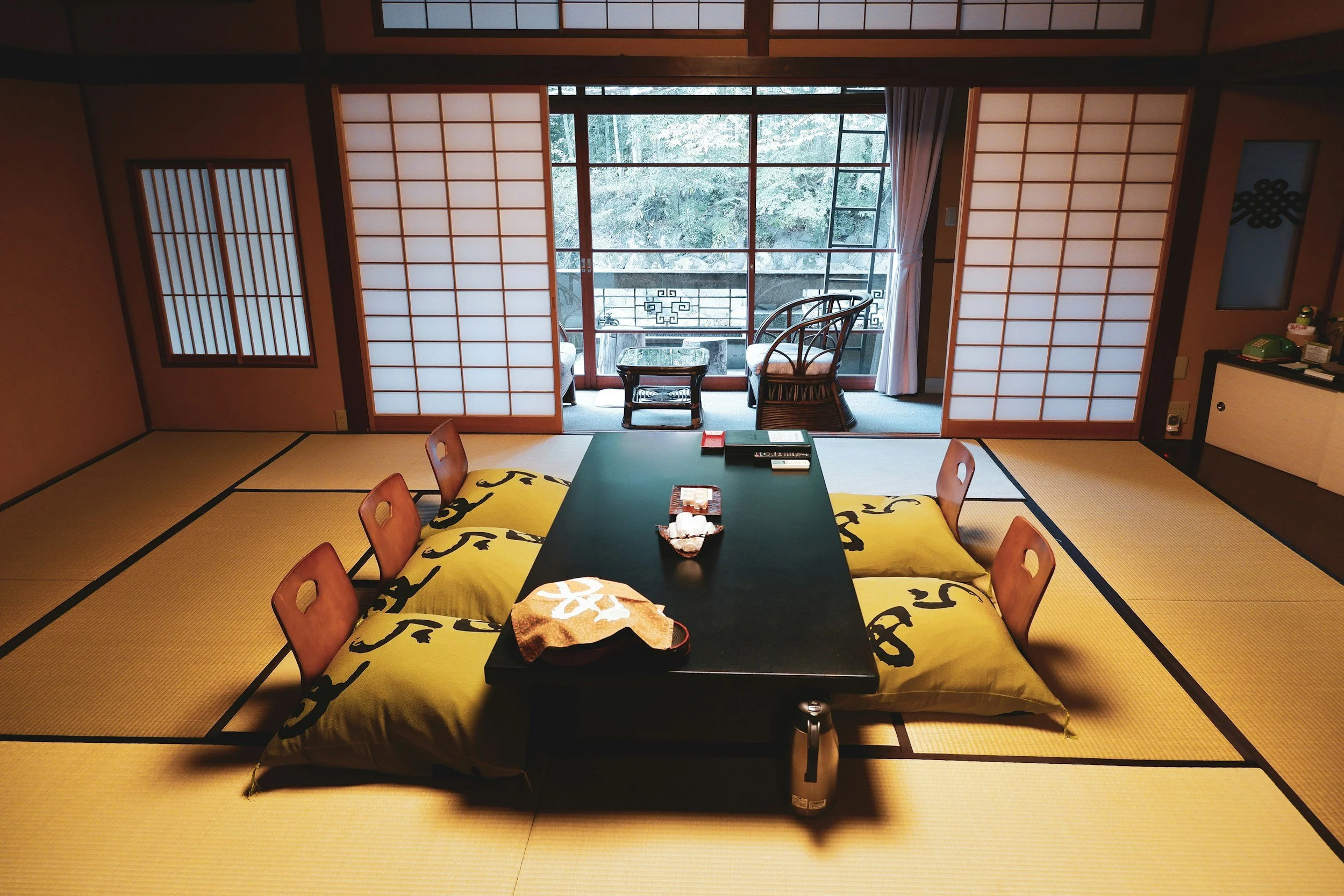 Traditional Japanese tatami room with a low black table, four floor cushions with yellow covers, shoji sliding doors, a sitting area with two chairs and a table on a balcony, and a view of greenery outside.