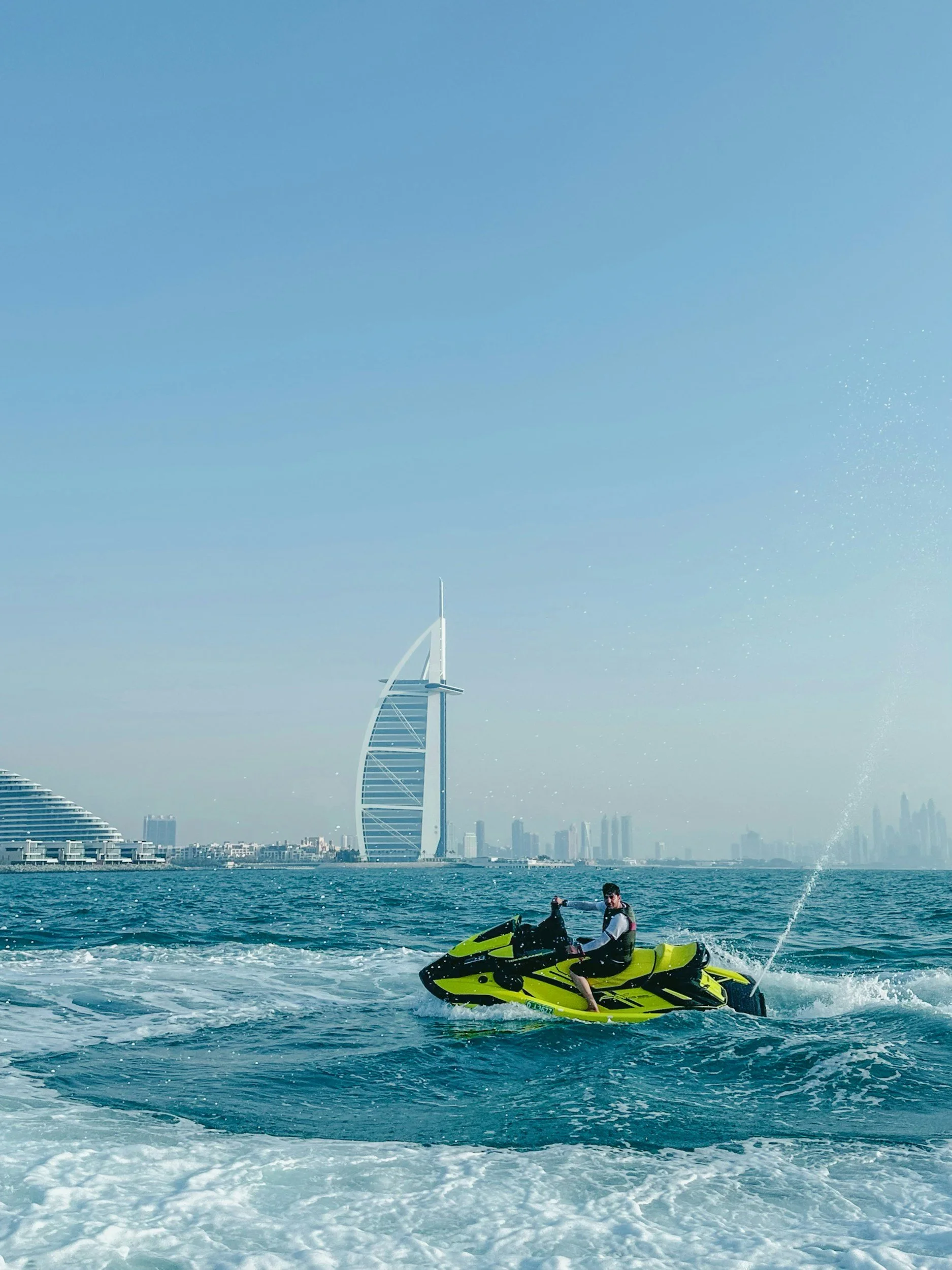 A man riding a yellow jet ski on the water with the Dubai skyline, including the Burj Al Arab hotel, in the background.