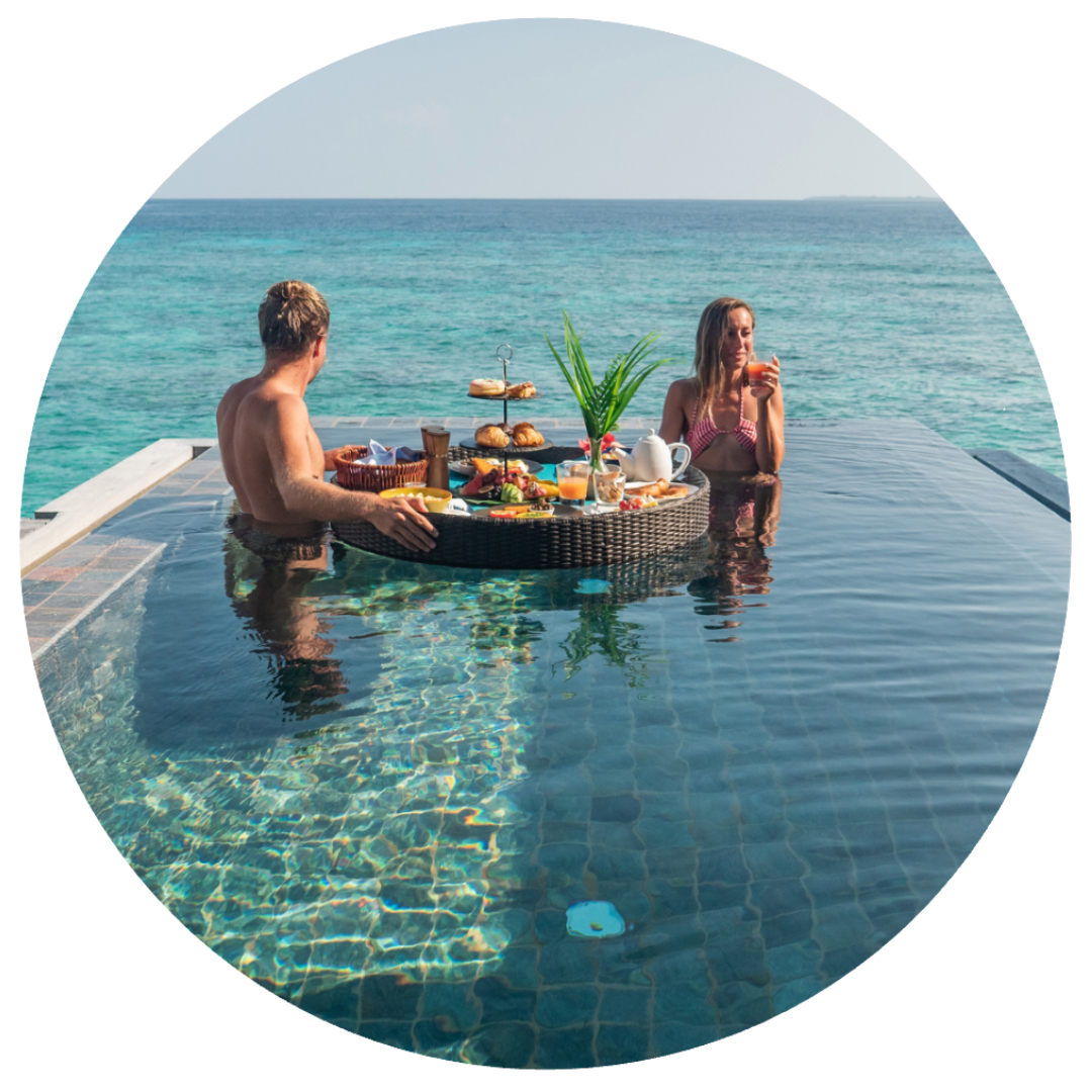 Two people enjoying breakfast in an infinity pool overlooking the ocean, with a floating tray of food including pastries, fruit, and drinks.