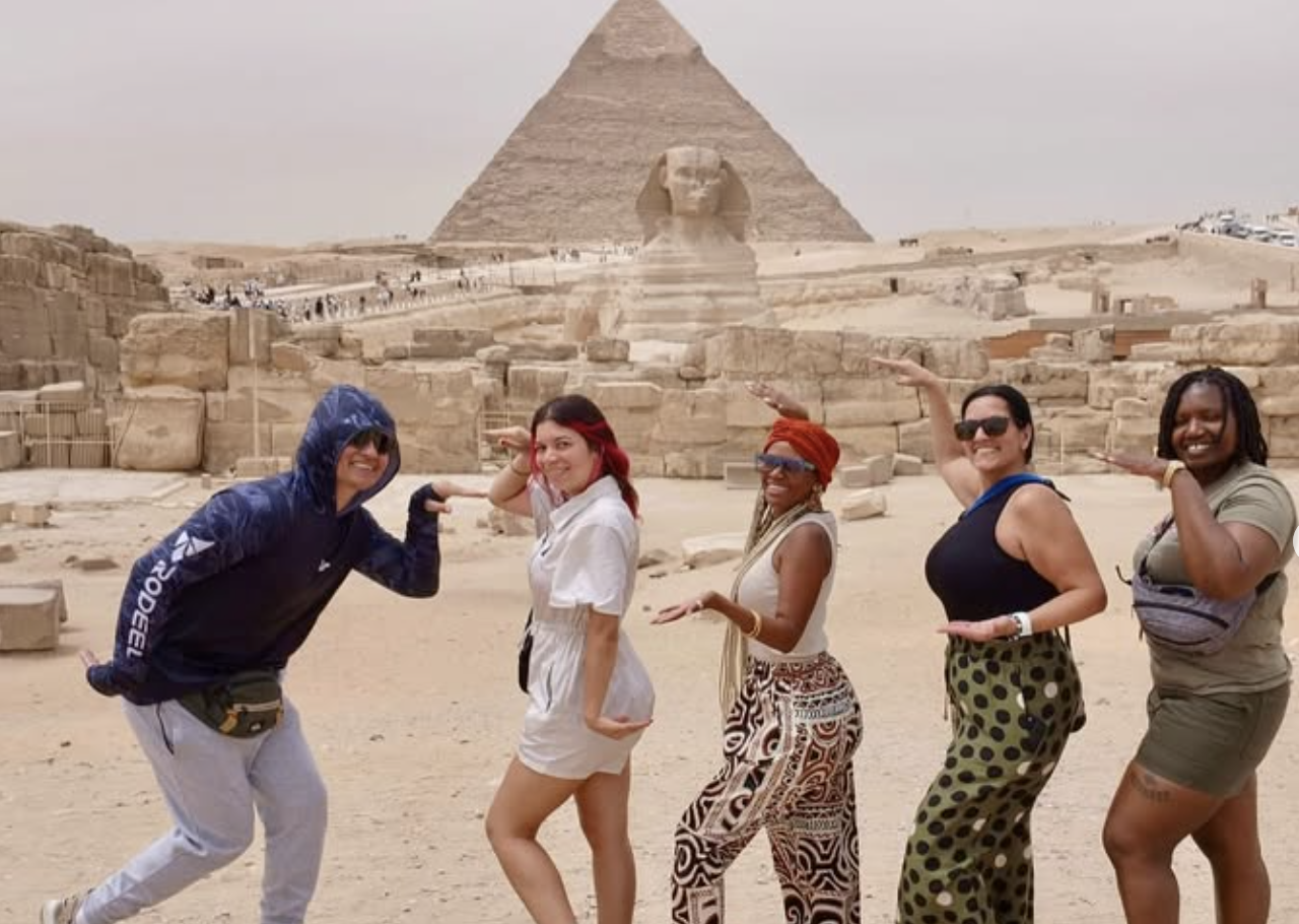 Five women posing in front of the pyramids and the Sphinx in Egypt, with desert landscape in the background.