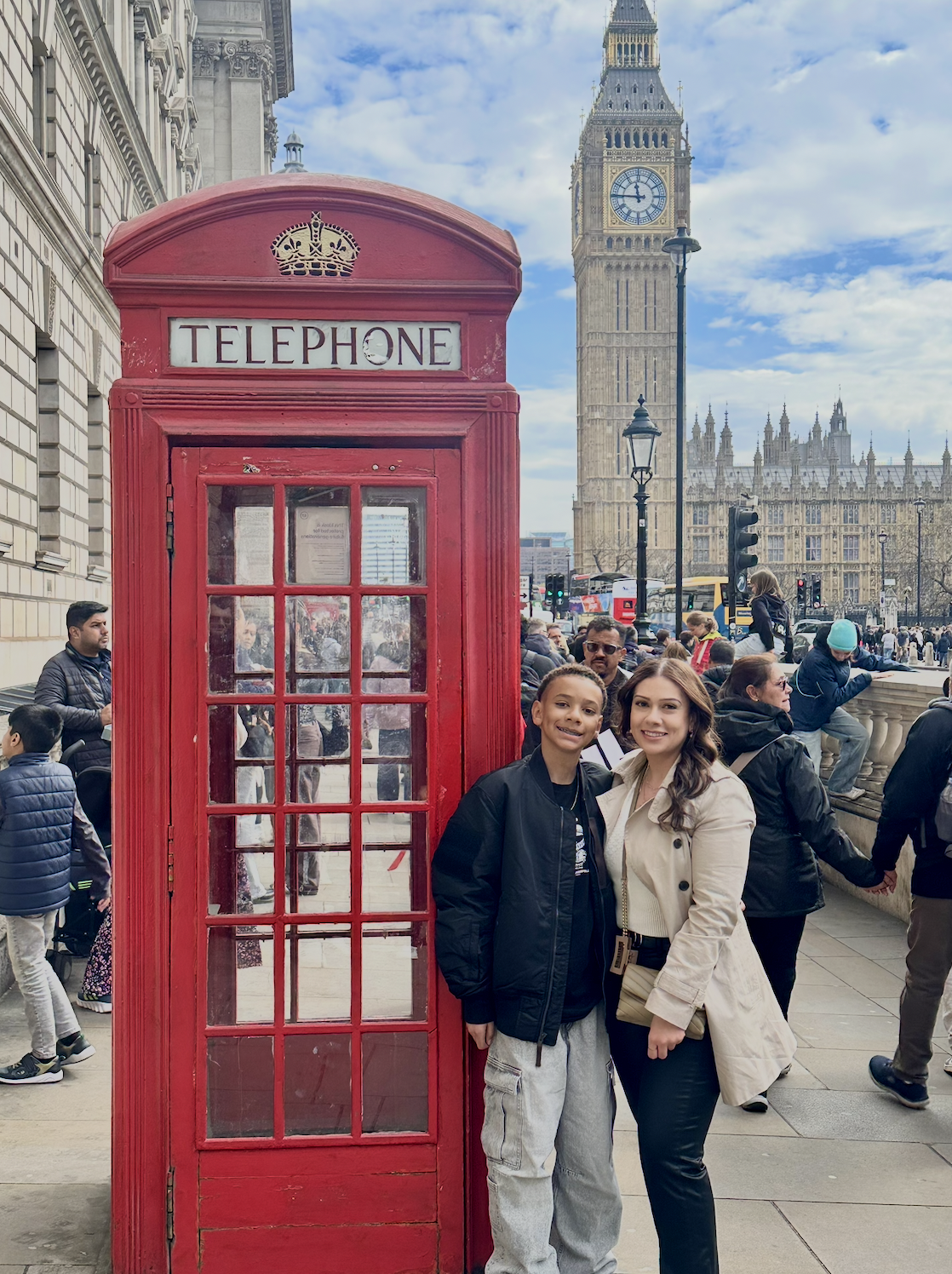 Two women stand smiling in front of a classic red British telephone booth with Big Ben clock tower in the background. The scene is busy with many people walking along a city street, with some seated on a ledge and others crossing the street.