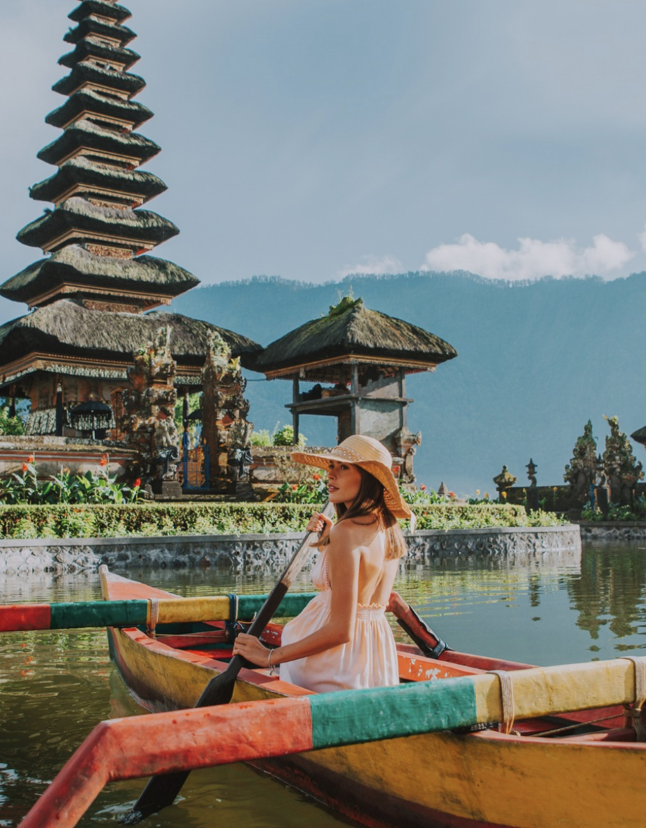 A woman sitting in a colorful kayak on a lake with traditional Balinese temples and mountains in the background.