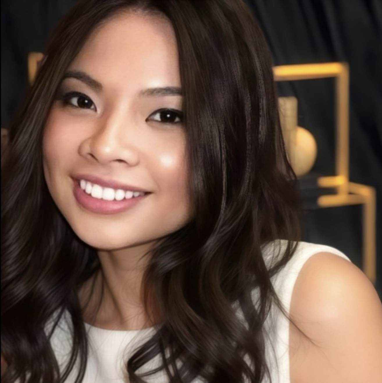 Close-up of a young woman with long, wavy dark hair, smiling and looking at the camera, with a modern dark interior background.