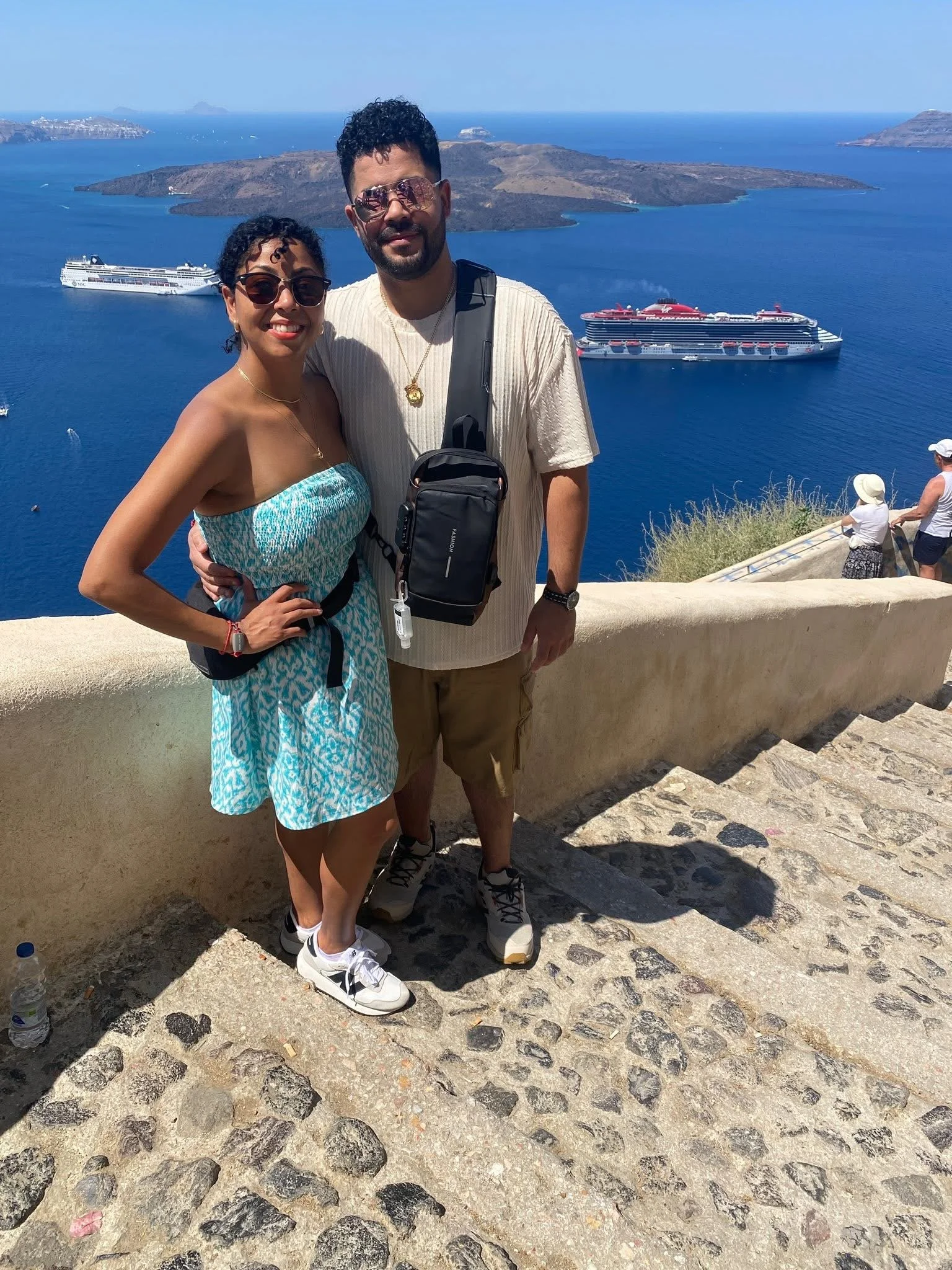 A couple posing on a stone pathway overlooking the ocean with large ships and volcanic islands in the background, likely in Santorini, Greece.
