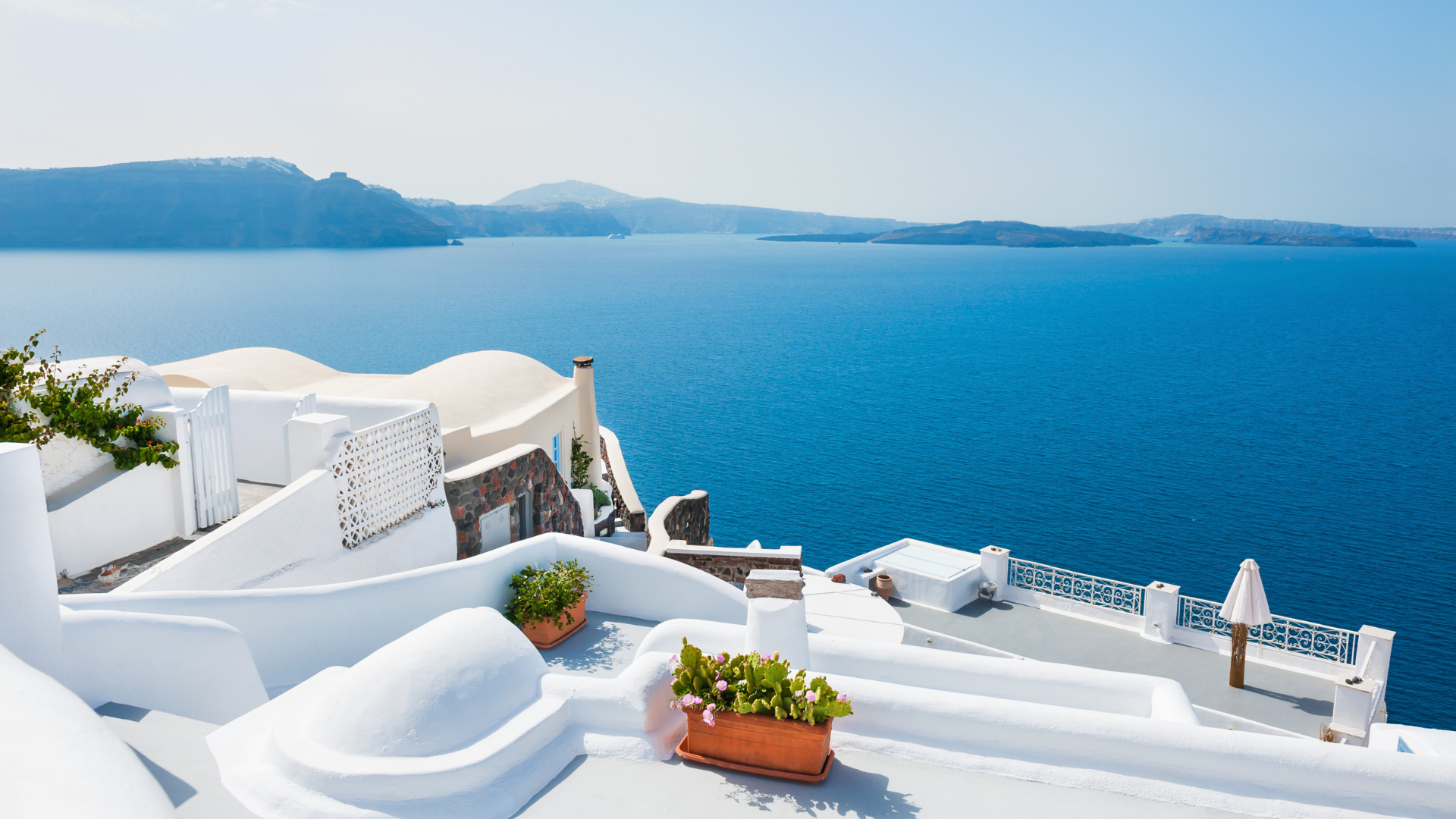 White buildings with curved roofs and potted plants overlooking deep blue sea and distant islands.
