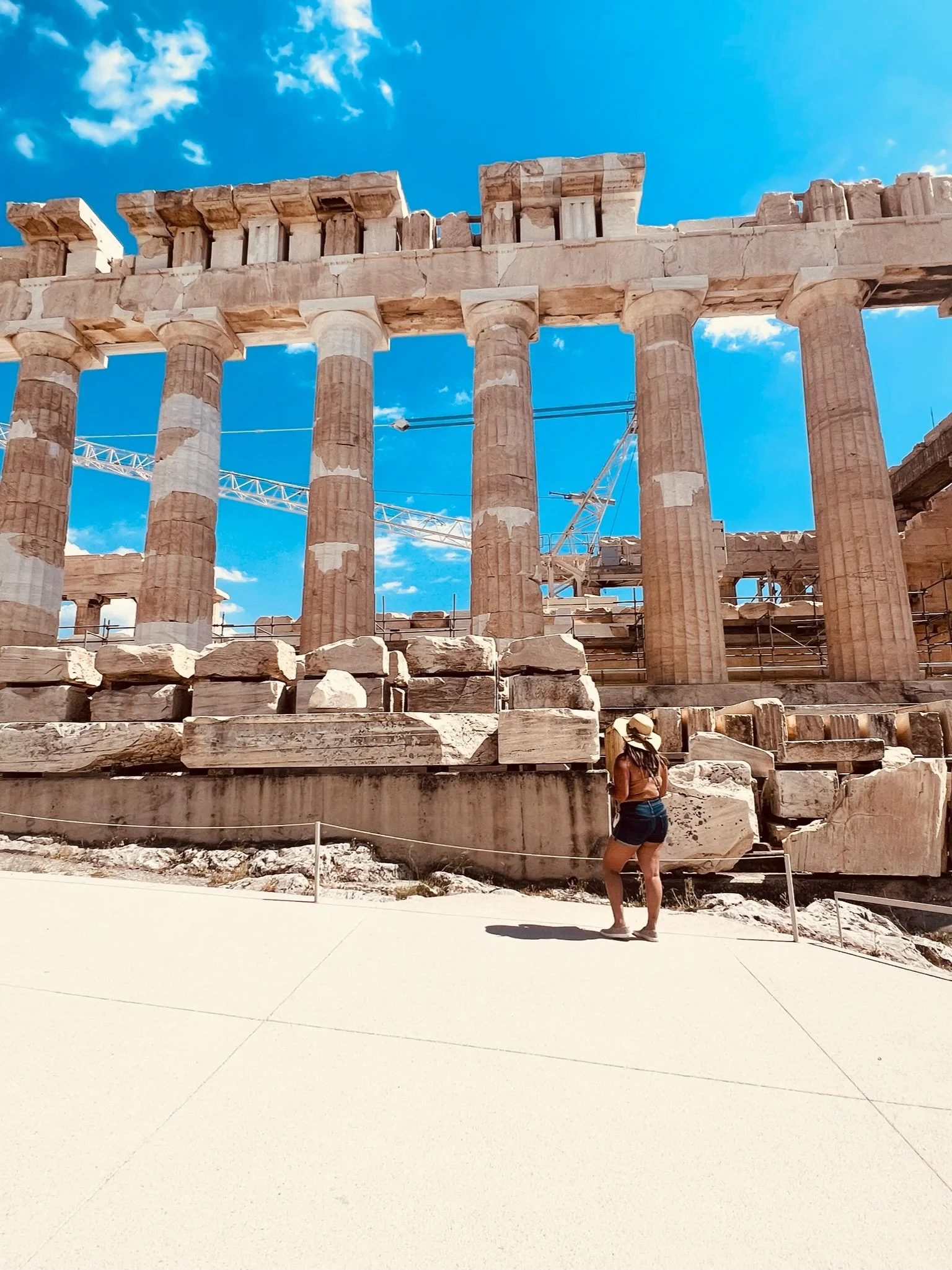 A woman standing in front of ancient Greek ruins with tall columns, wearing a hat, tank top, and shorts, under a bright blue sky with scattered clouds.