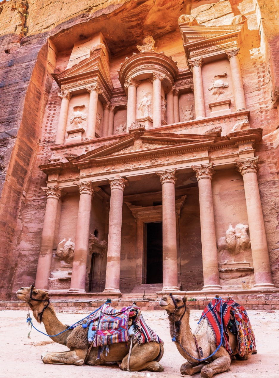 Two camels with colorful blankets and saddles resting in front of the ancient rock-cut city of Petra, Jordan with its intricate stone facade and columns carved into the pink sandstone cliffs.