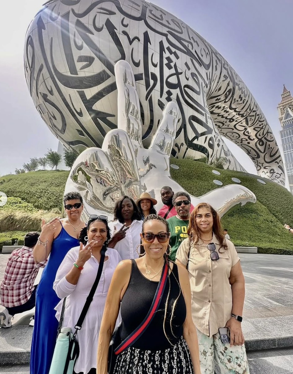 A group of people taking a selfie in front of a large, artistic sculpture of a hand making a peace sign. The sculpture features calligraphy-style patterns and is set outdoors with greenery and a tall building in the background.