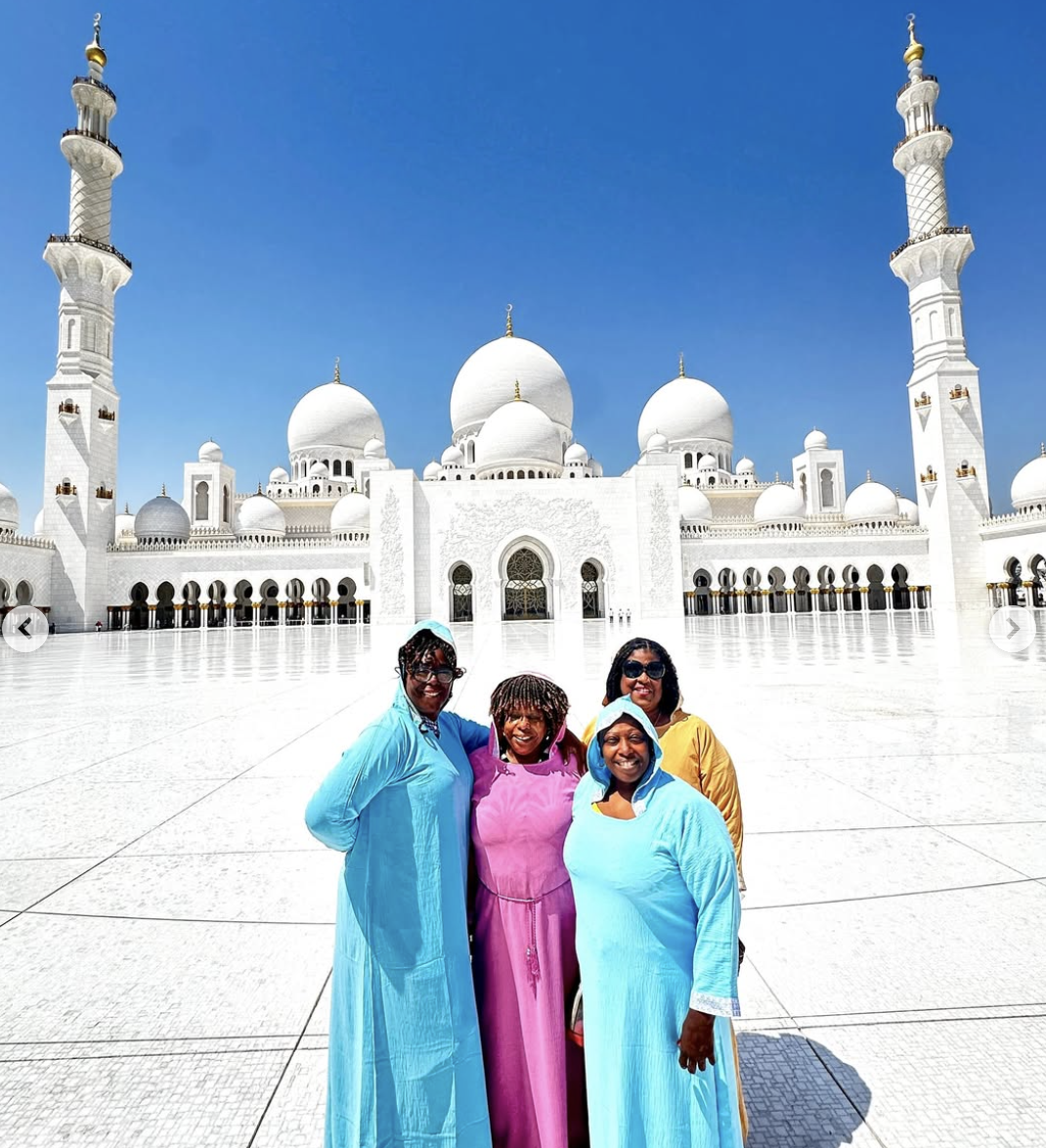 Four women in colorful dresses standing in front of the white marble architecture of the Sheikh Zayed Grand Mosque, with its domes and minarets under a clear blue sky.