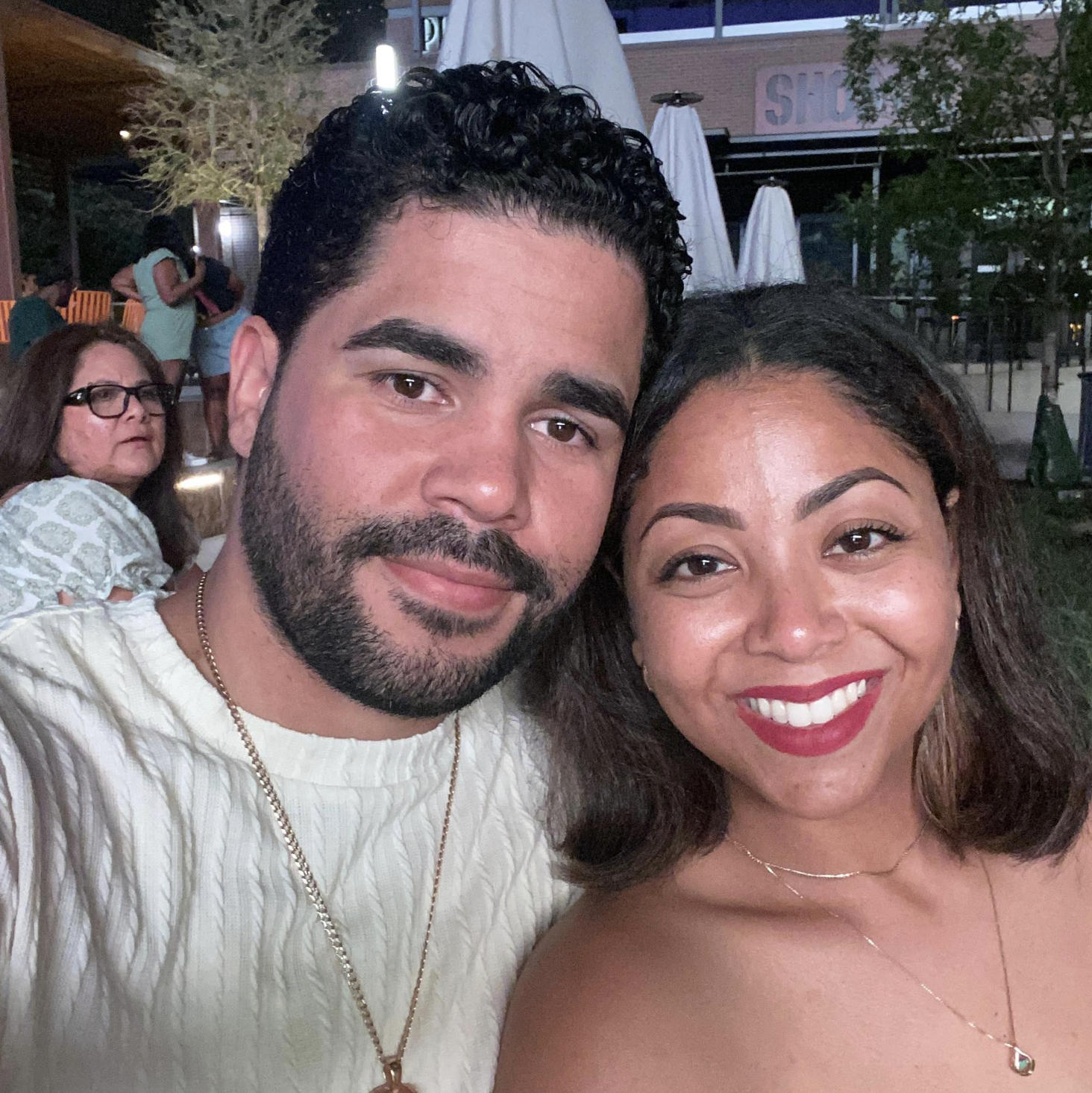 A smiling man and woman taking a selfie at an outdoor event during evening. The man has dark curly hair and a beard, wearing a cream-colored sweater and gold chain. The woman has dark hair, wearing red lipstick, necklace, and strapless top. In the background, other people and outdoor seating are visible.