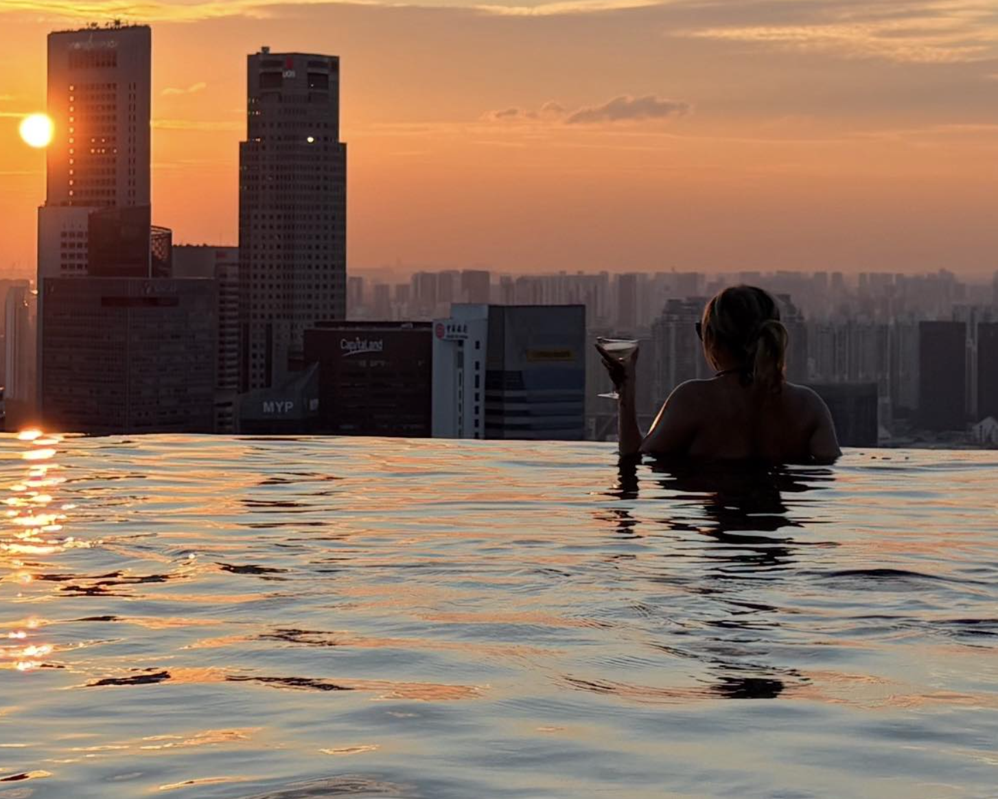 A woman in an infinity pool holding a martini glass during sunset with an urban city skyline in the background.