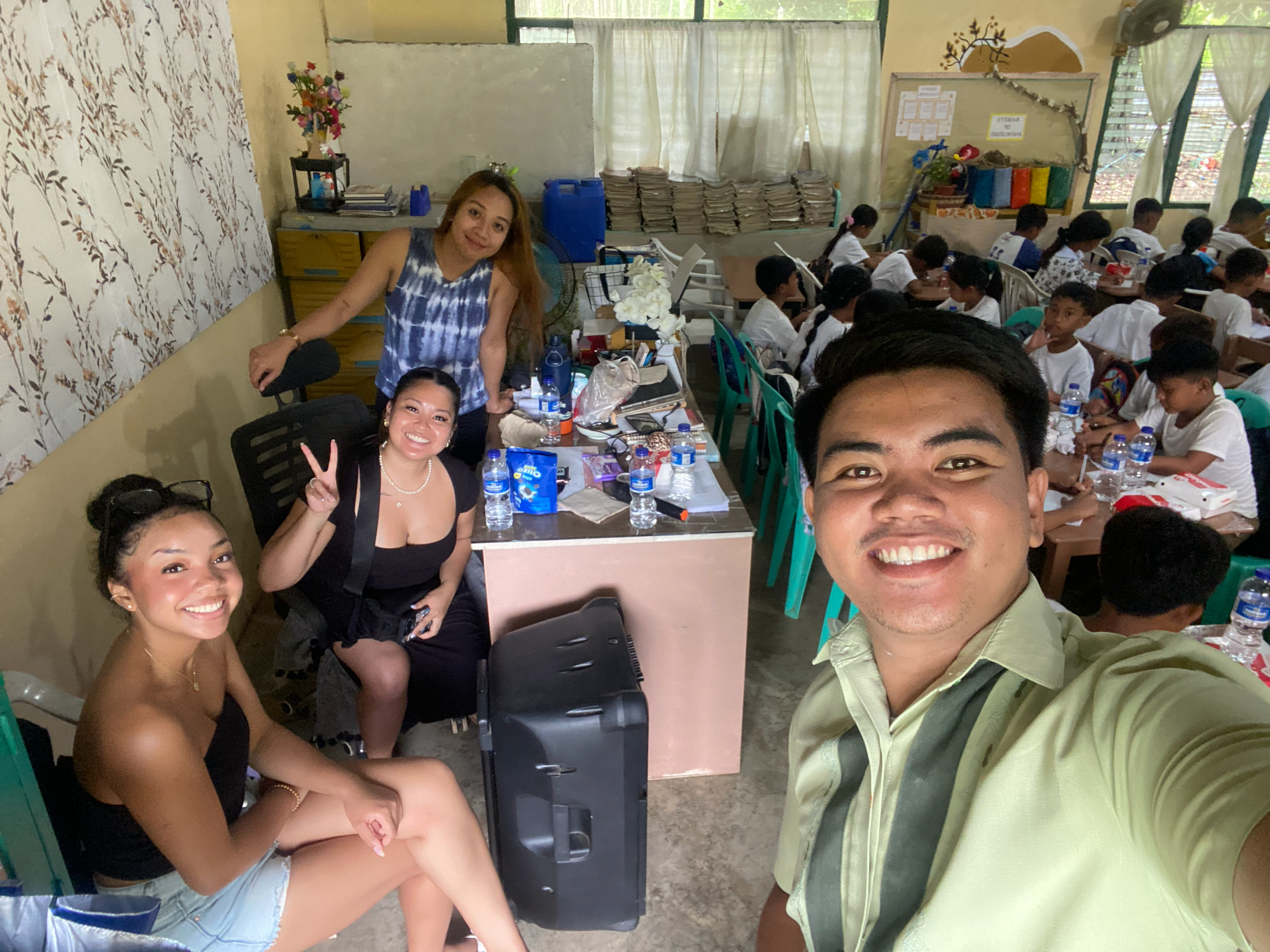 Group of four people smiling and taking a selfie in a classroom with students seated at desks in the background. The group includes three women and one man, with various items and water bottles on a nearby table.