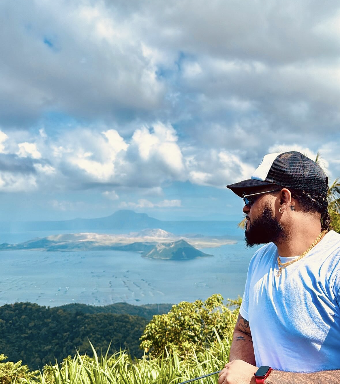 A man with sunglasses, a black and white cap, a white t-shirt, gold chains, and a red smartwatch looking at a distant volcano and cove with mountains in the background.