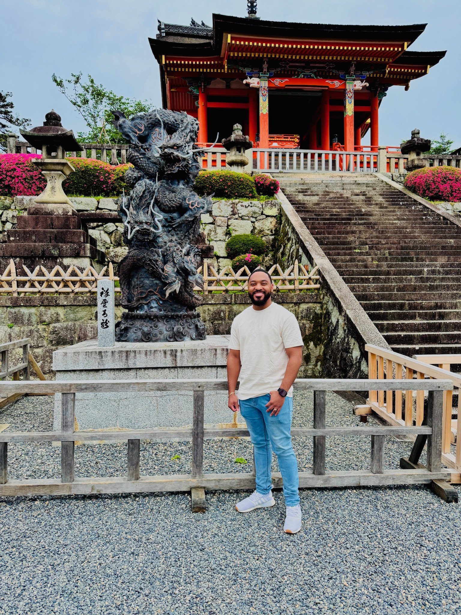 A smiling man in a beige t-shirt, blue jeans, and white sneakers standing in front of a Japanese-style temple with a dragon statue and stone stairs.