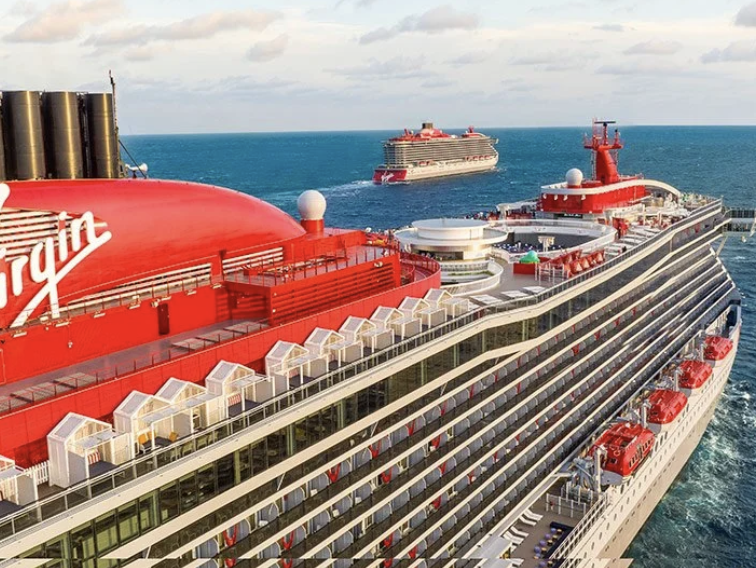 Aerial view of a large red Virgin cruise ship with multiple decks, lifeboats, and outdoor lounge areas, with two other cruise ships sailing in the ocean in the background.