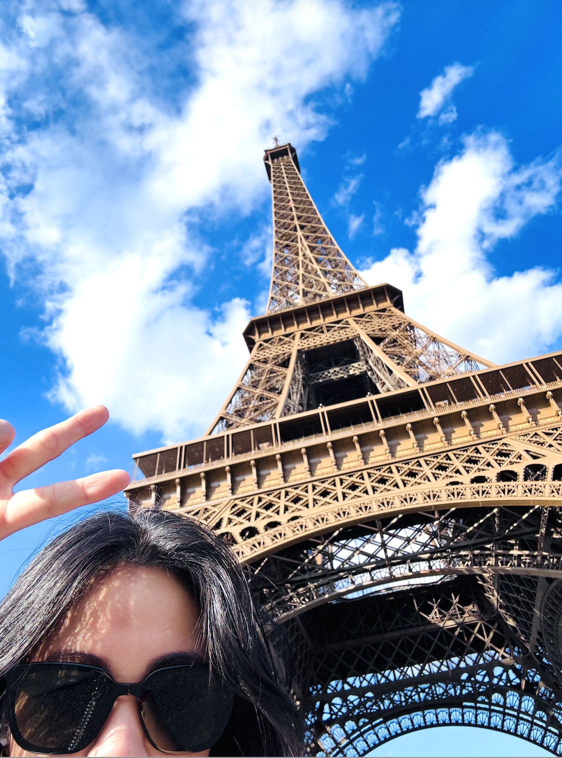 Close-up of a woman's face wearing sunglasses, with the Eiffel Tower behind her against a blue sky with clouds.