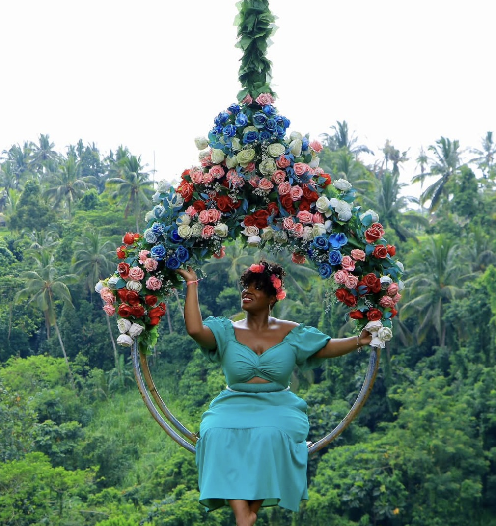 A woman in a turquoise dress sitting on a floral swing with a floral canopy, surrounded by lush green trees.