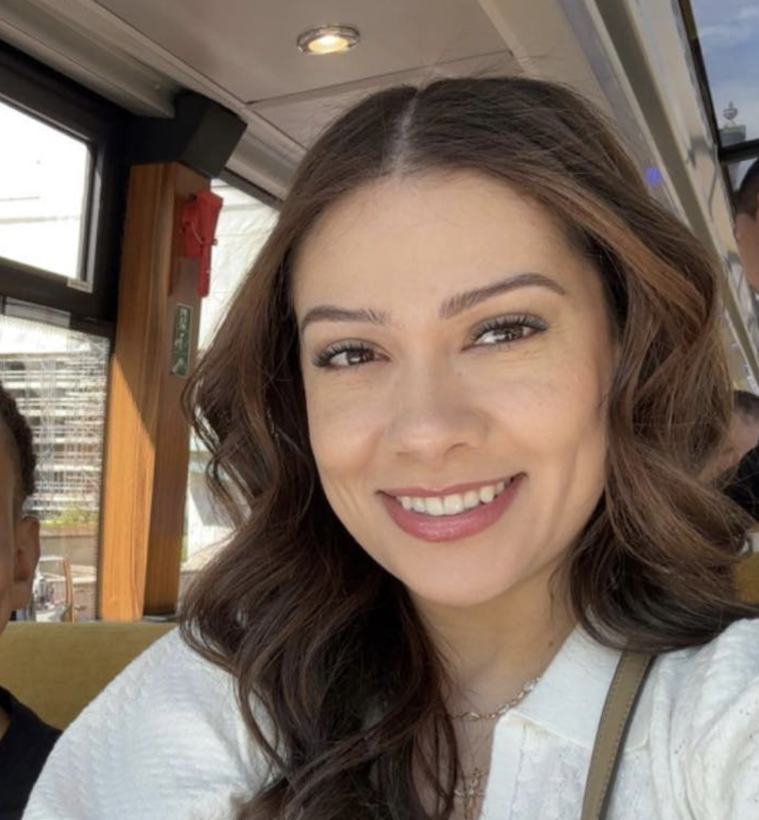 A woman with wavy brown hair smiling at the camera on a bus or train.