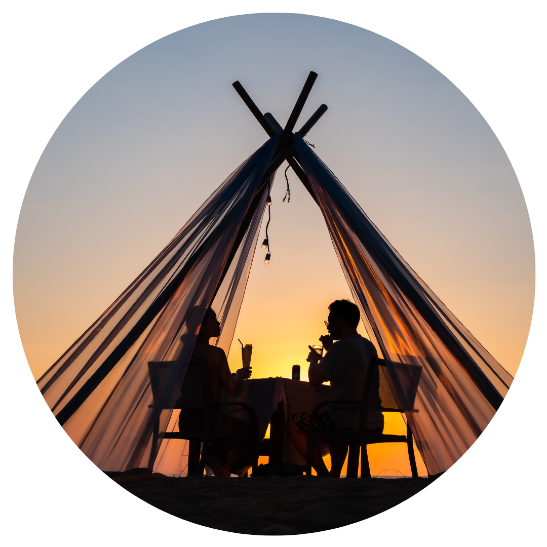 Silhouettes of two people sitting at a table inside a tent during sunset, drinking beverages. The scene has a beach vibe with a warm, colorful sky.
