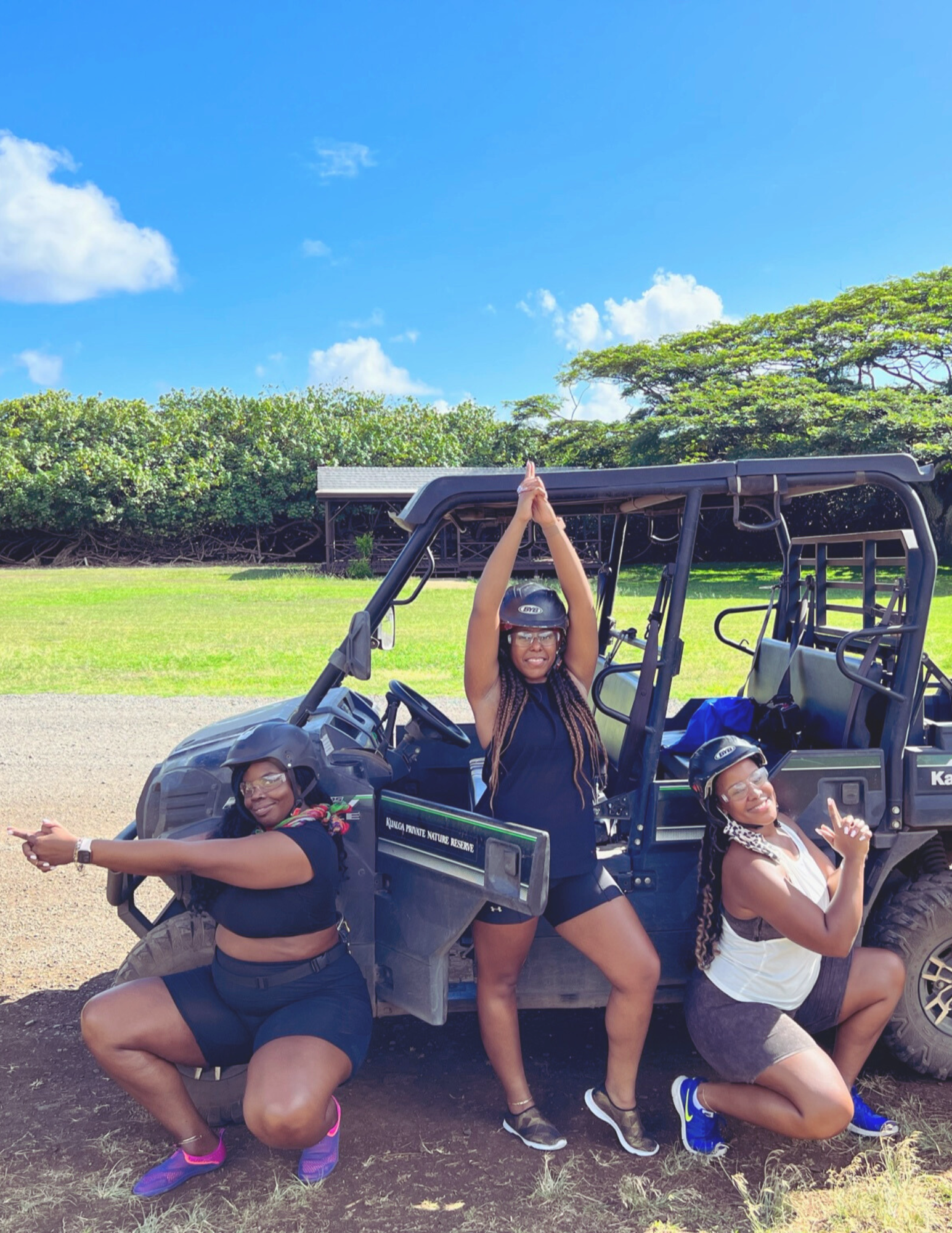 Three women wearing helmets and glasses posing with a black off-road vehicle in a green outdoor setting under a blue sky with some clouds.