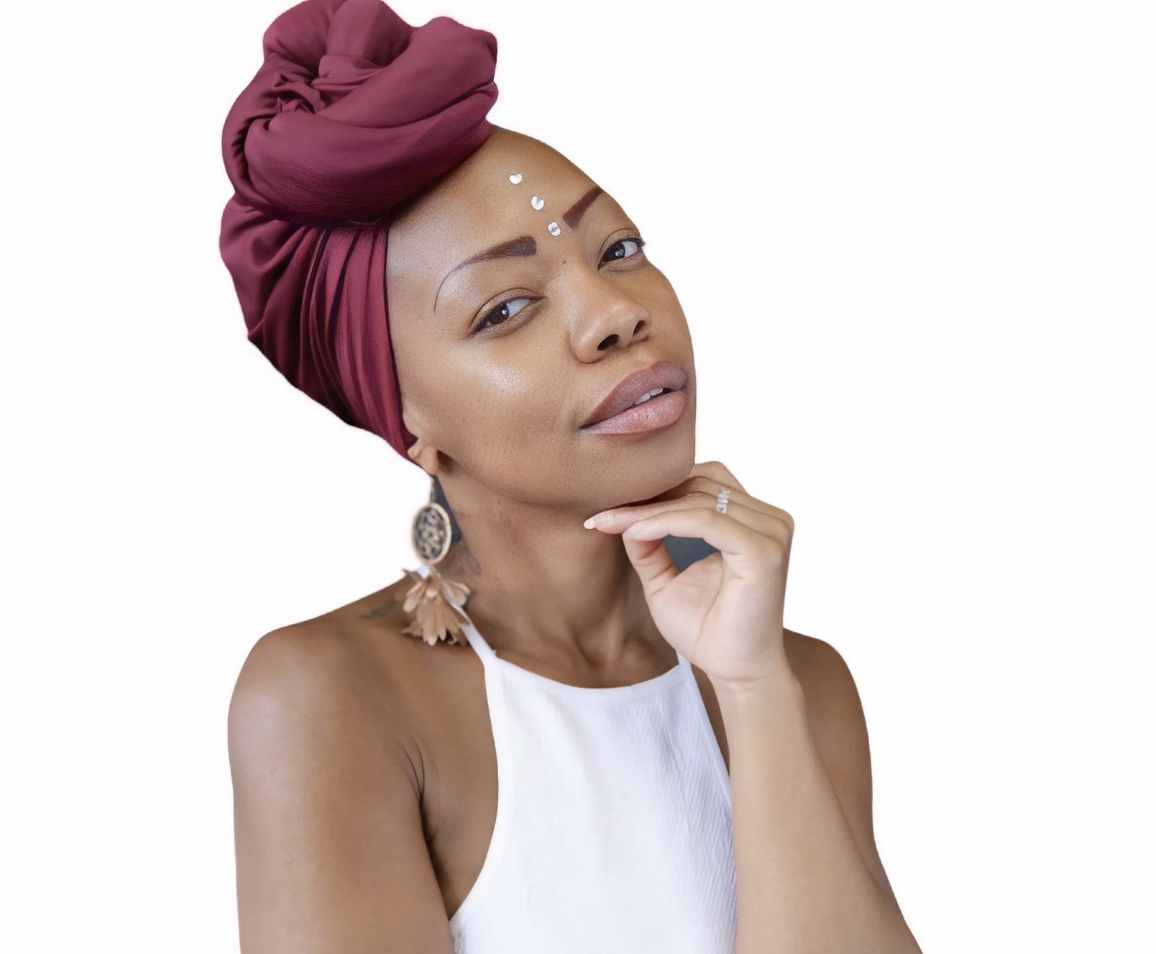 A woman wearing a large maroon headwrap, white top, and floral earrings, with jewelry on her face, posing with her hand near her chin against a white background.