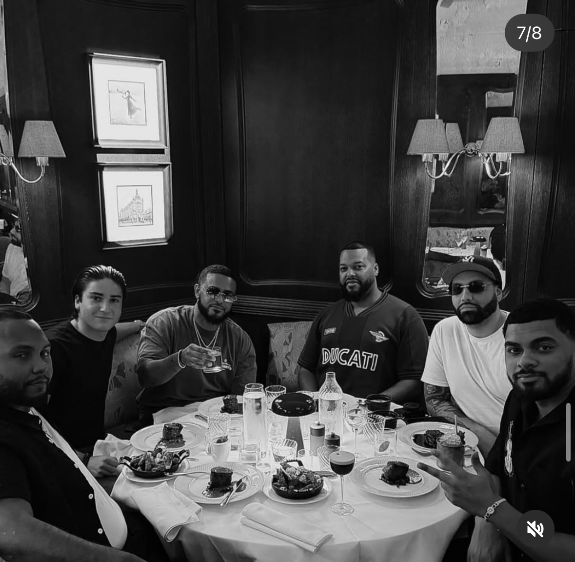 Six people sitting around a dining table with food and drinks at a restaurant. The setting features wood-paneled walls, wall-mounted lamps, and framed artwork. Some individuals are wearing sunglasses and casual clothing.