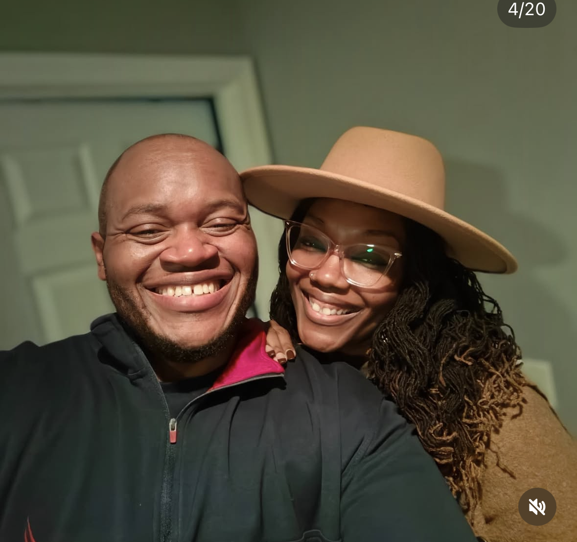 A smiling man and woman taking a selfie together indoors. The man has a shaved head and is wearing a black jacket with a pink collar. The woman has long curly hair, is wearing glasses and a wide-brimmed tan hat, and is dressed in a brown top.