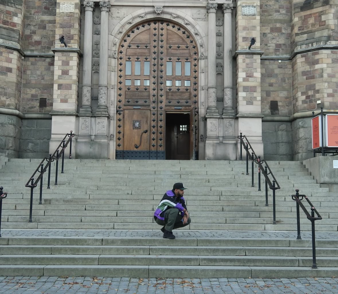 A man crouching on the steps in front of a large ornate wooden door of a historic stone building.
