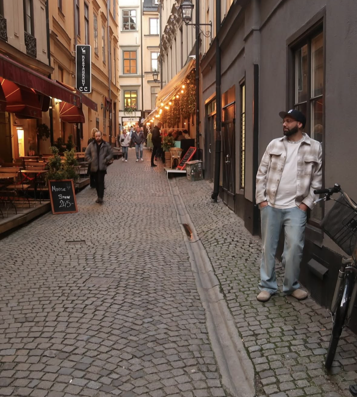 People walking along a cobblestone street with shops and outdoor dining. A man with a bicycle stands near a gray building on the right. The street is decorated with string lights, and there's a sign advertising moose stew.